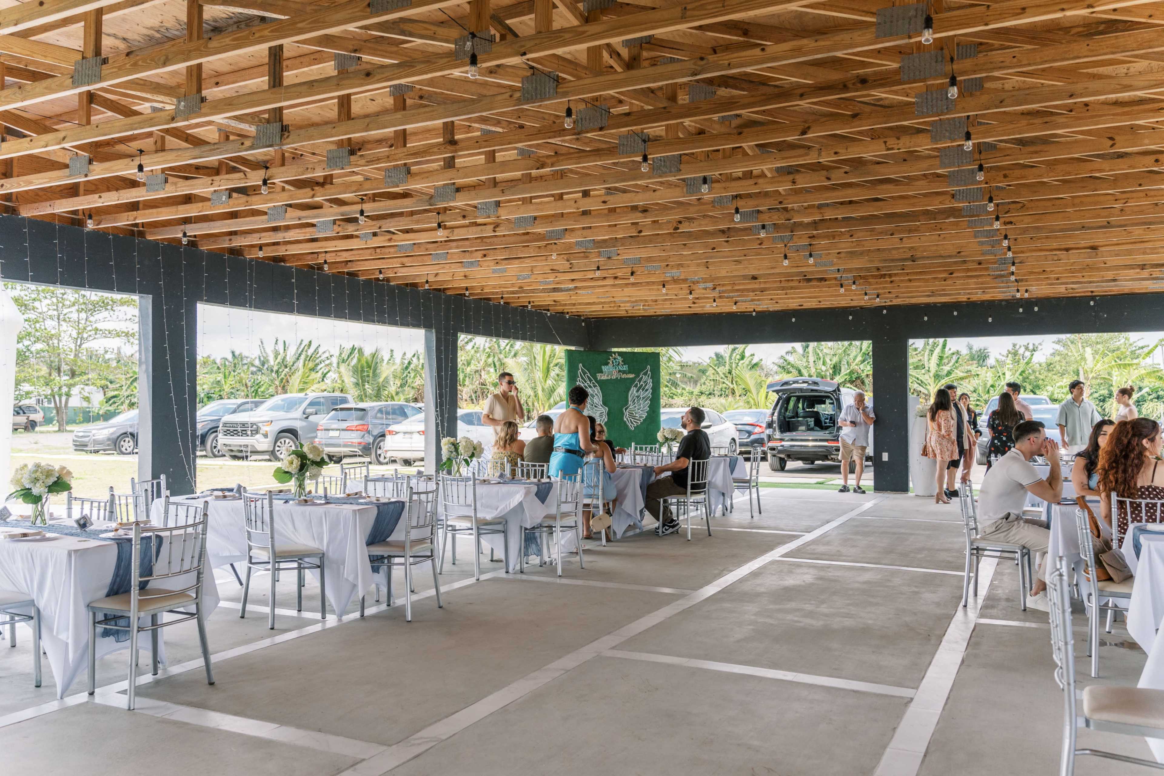 A spacious outdoor venue features decorated tables and guests mingling under a wooden ceiling with string lights, while cars are parked in the background.
