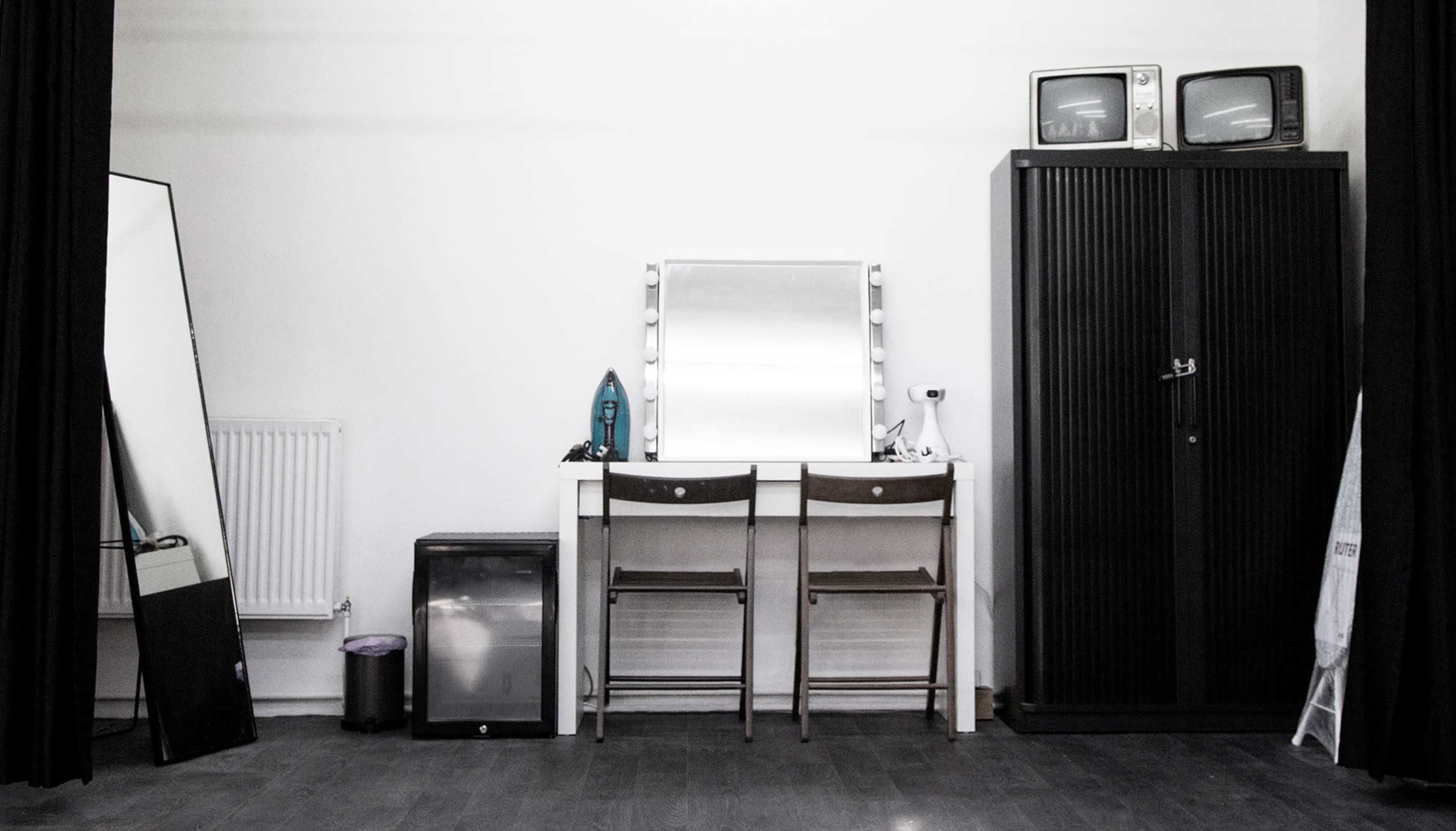 A minimalist room featuring a large mirror, two chairs, a small refrigerator, a pair of vintage TVs, and a tall cabinet against a white wall.