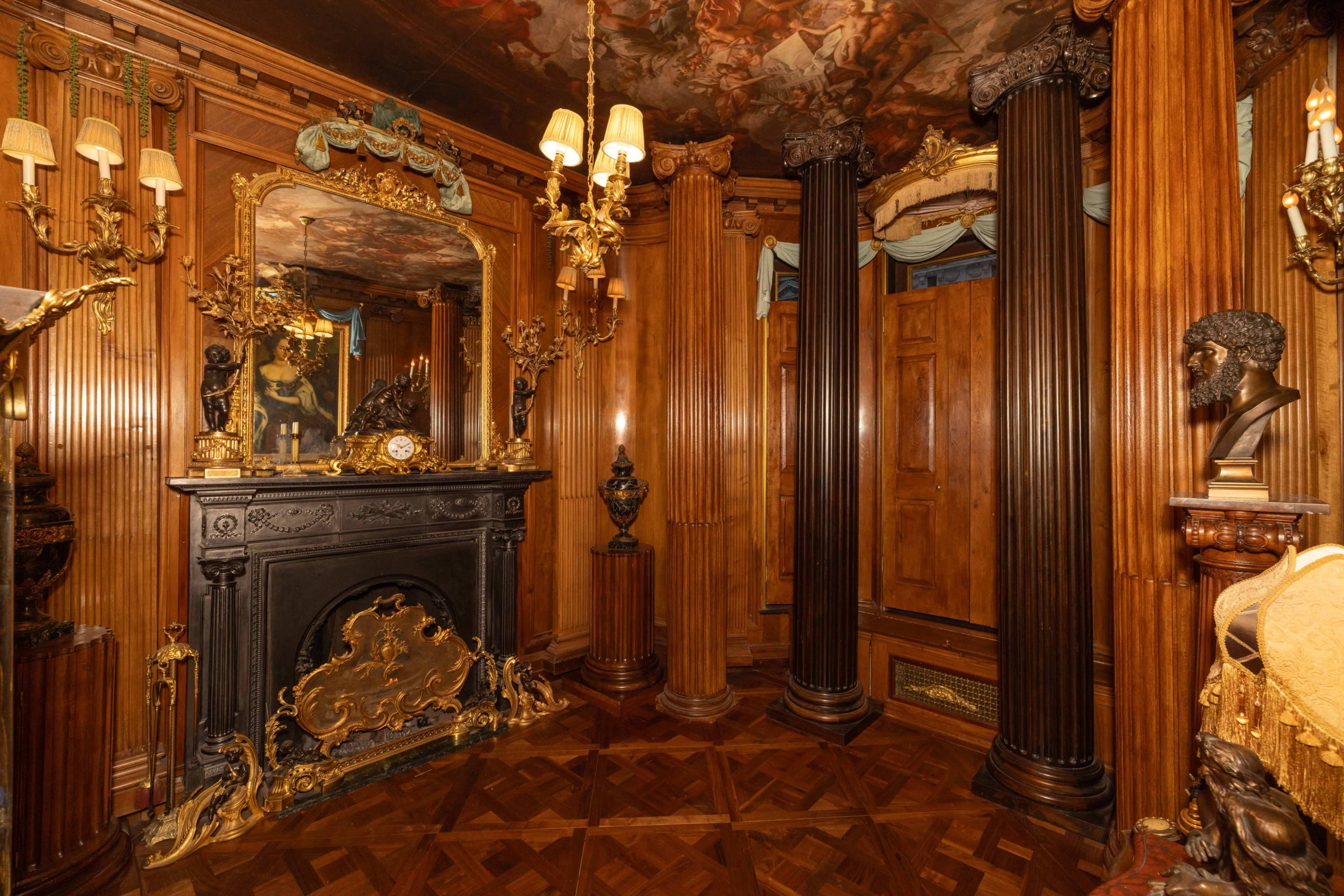 The image shows an ornate, richly decorated room featuring wooden pillars, a large fireplace with intricate design, and various decorative elements including sculptures and wall sconces.