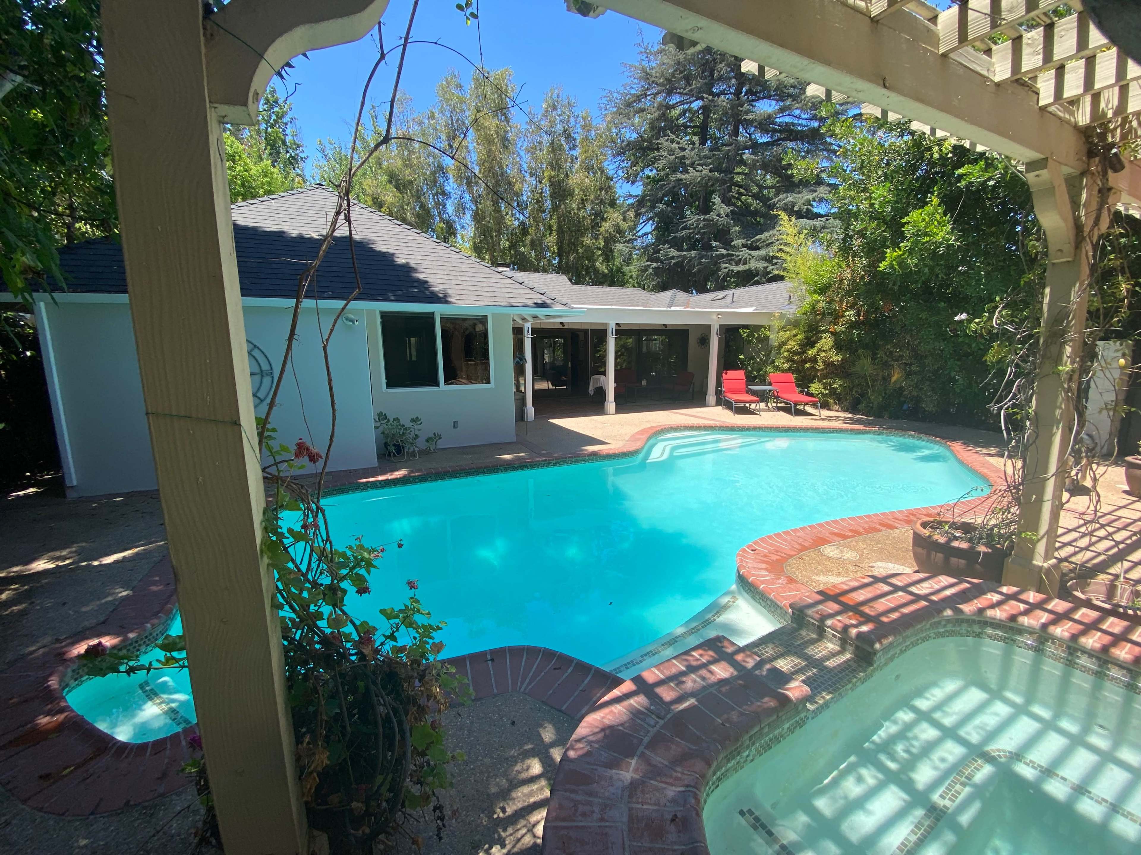 A residential backyard with a swimming pool, a spa, and lounge chairs, surrounded by greenery and a house in the background.