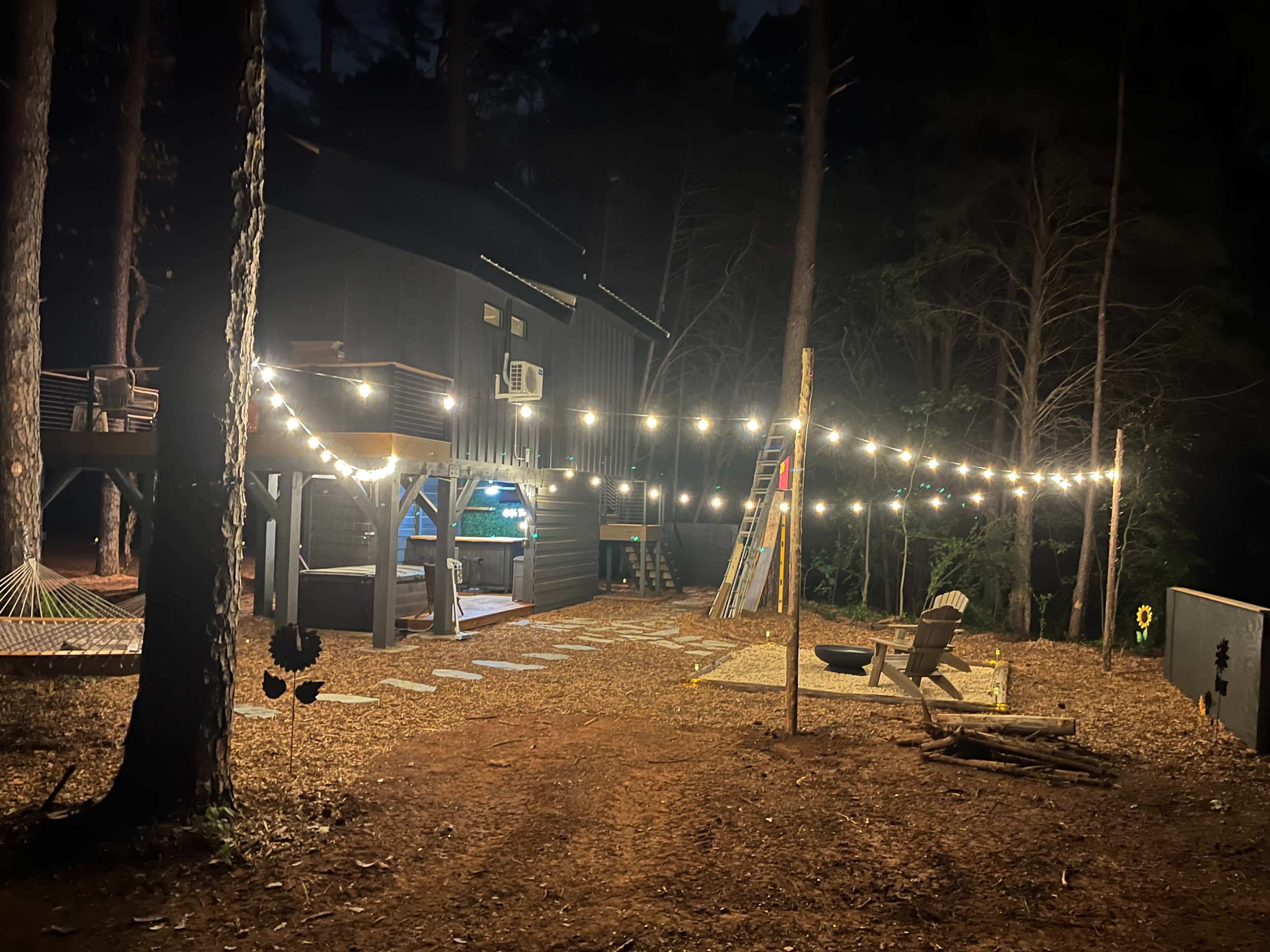 The image shows a dark outdoor area illuminated by string lights, with a wooden building, a hammock, and a fire pit surrounded by pine trees.