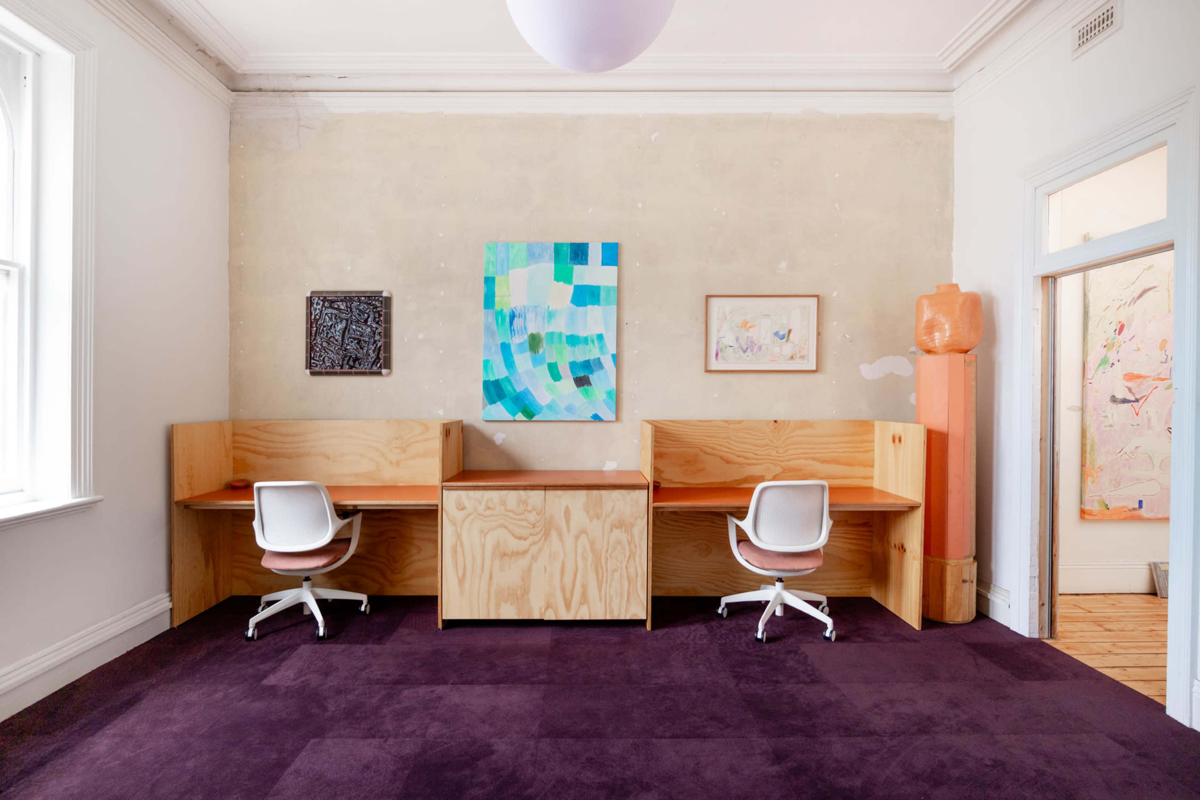 The image shows a spacious room featuring two wooden desks, each with a white chair, on a deep purple carpet, with contemporary artwork displayed on the walls.