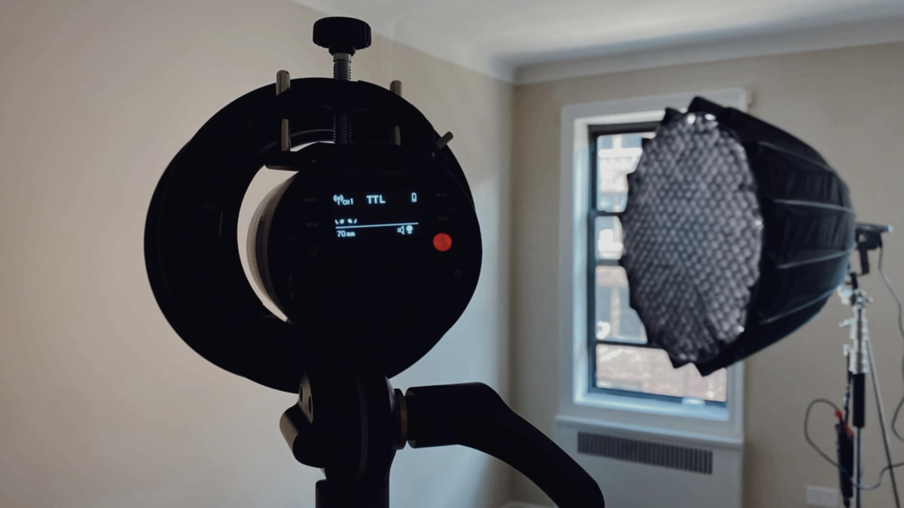 A camera flash device is set up in a well-lit room, with a softbox positioned nearby.