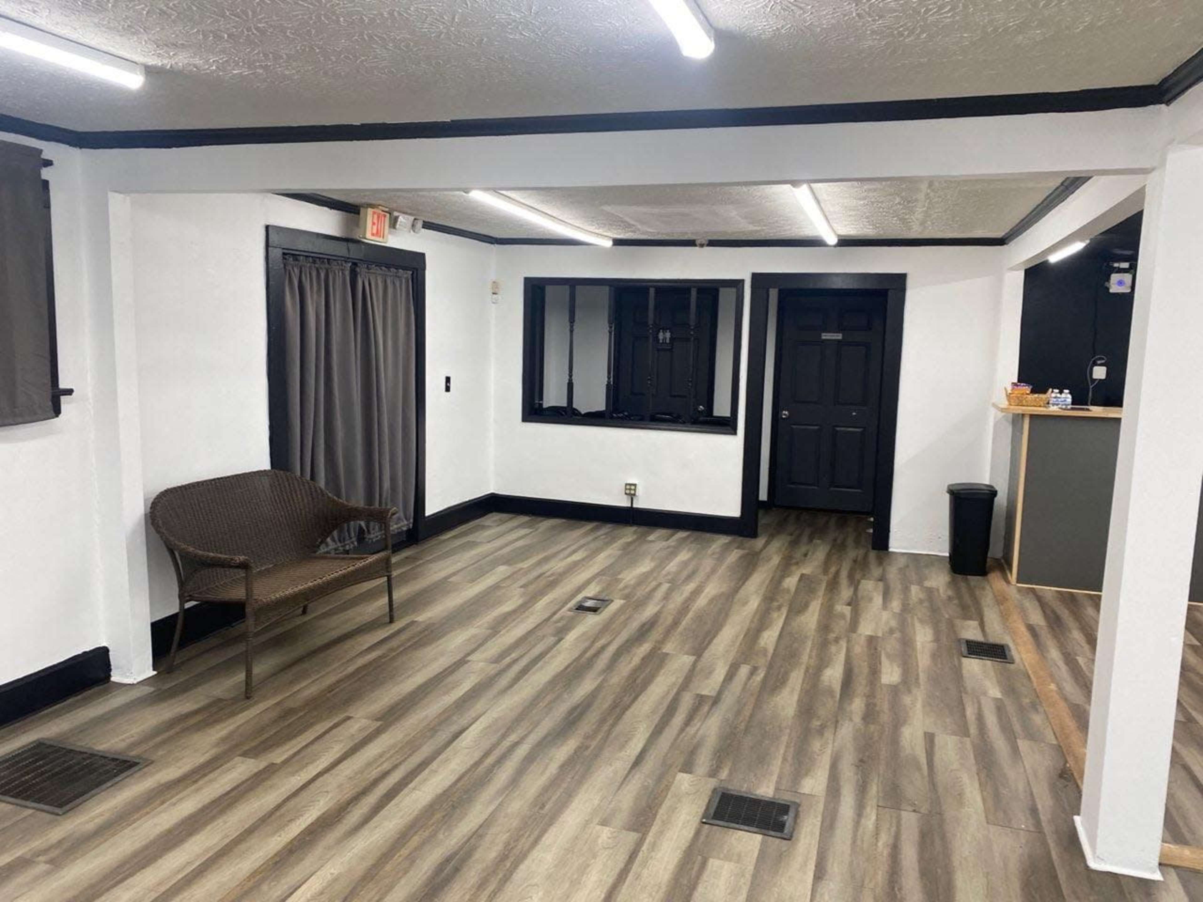 The image shows a sparsely furnished interior space with wood-like flooring, white walls, and black trim, featuring a chair, window, and an open doorway.