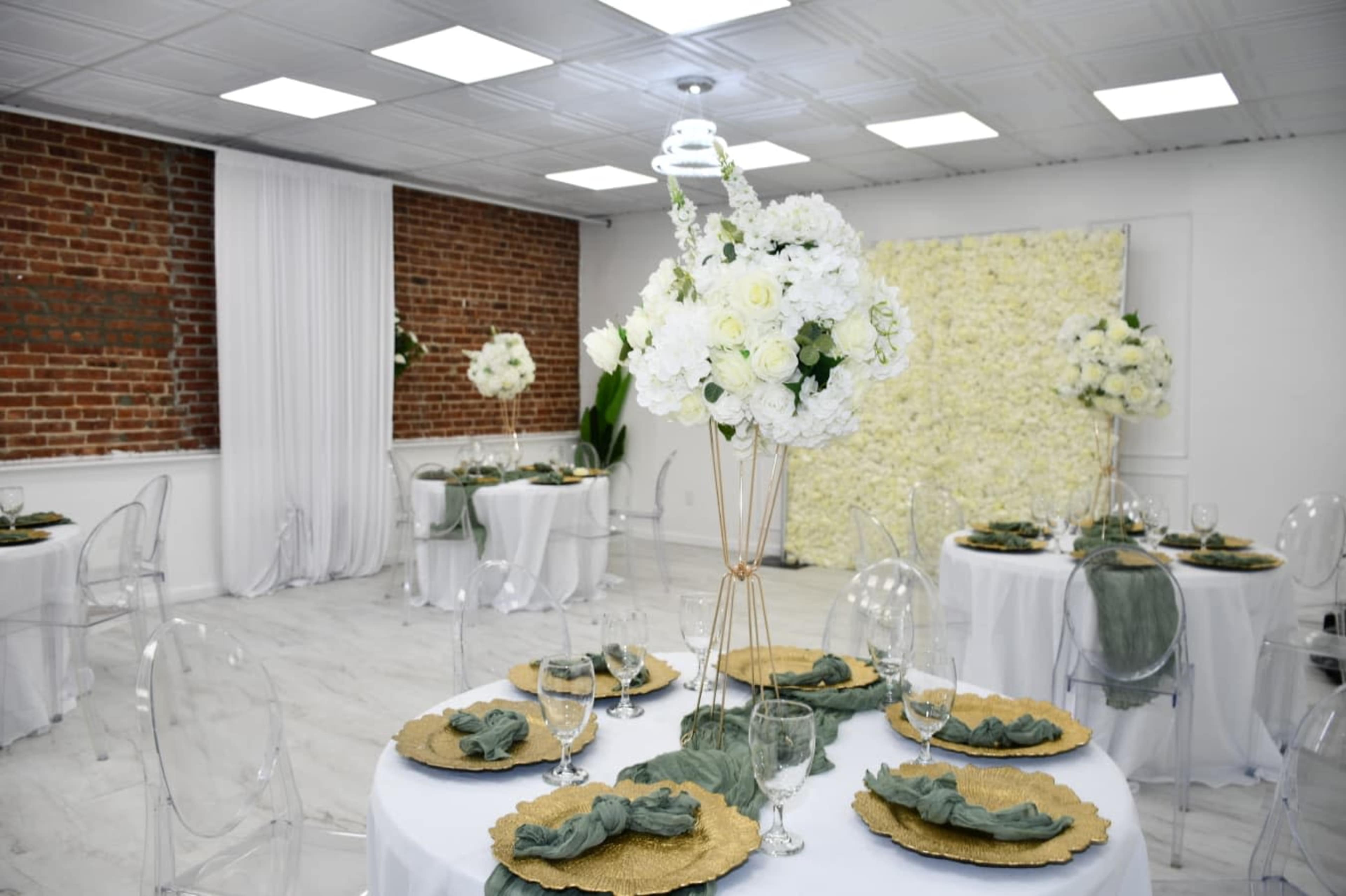 The elegantly designed event space features round tables with gold chargers, floral centerpieces, and a white flower wall backdrop against exposed brick.