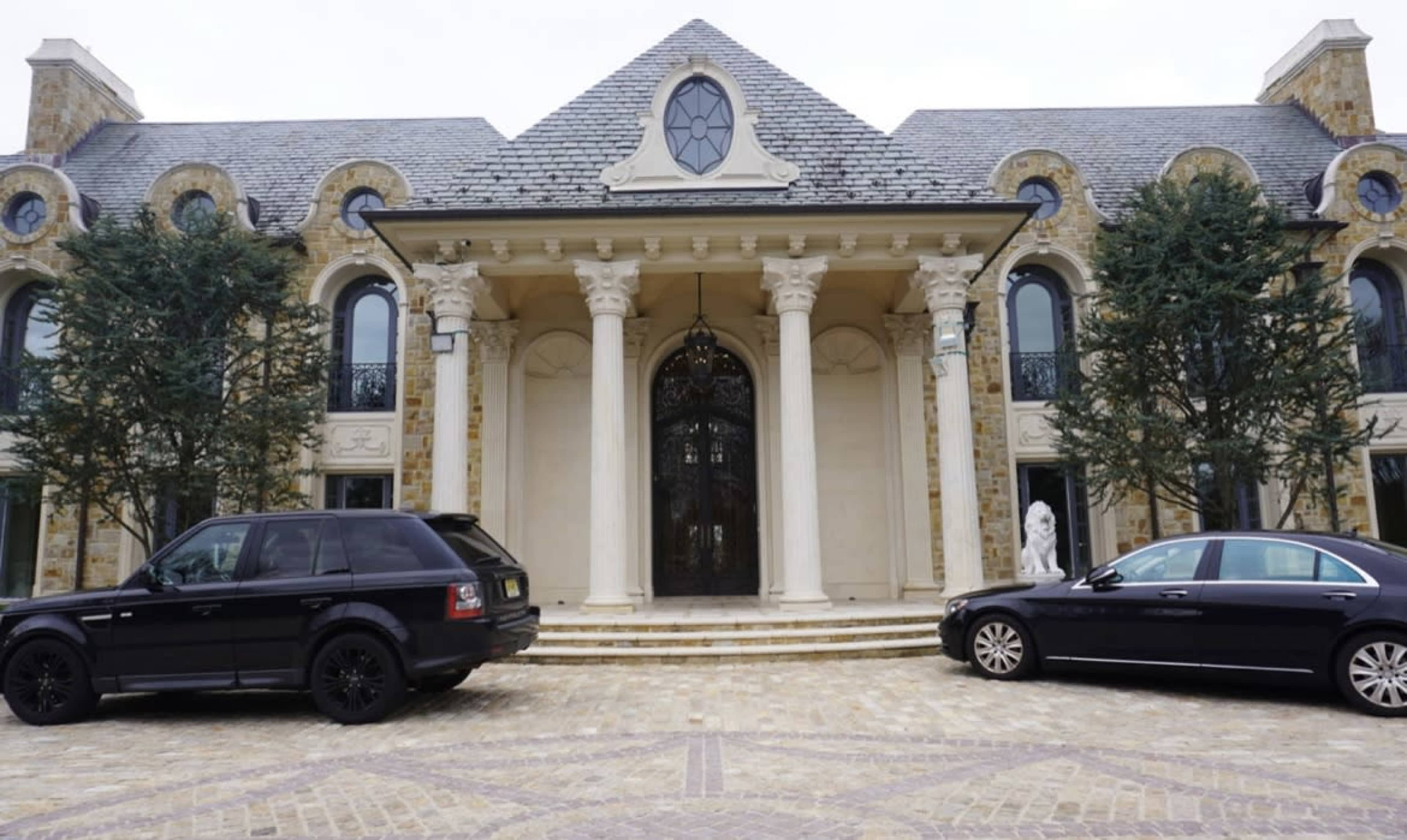 A large mansion with a symmetrical facade and ornate columns is flanked by two luxury cars parked on a cobblestone driveway.