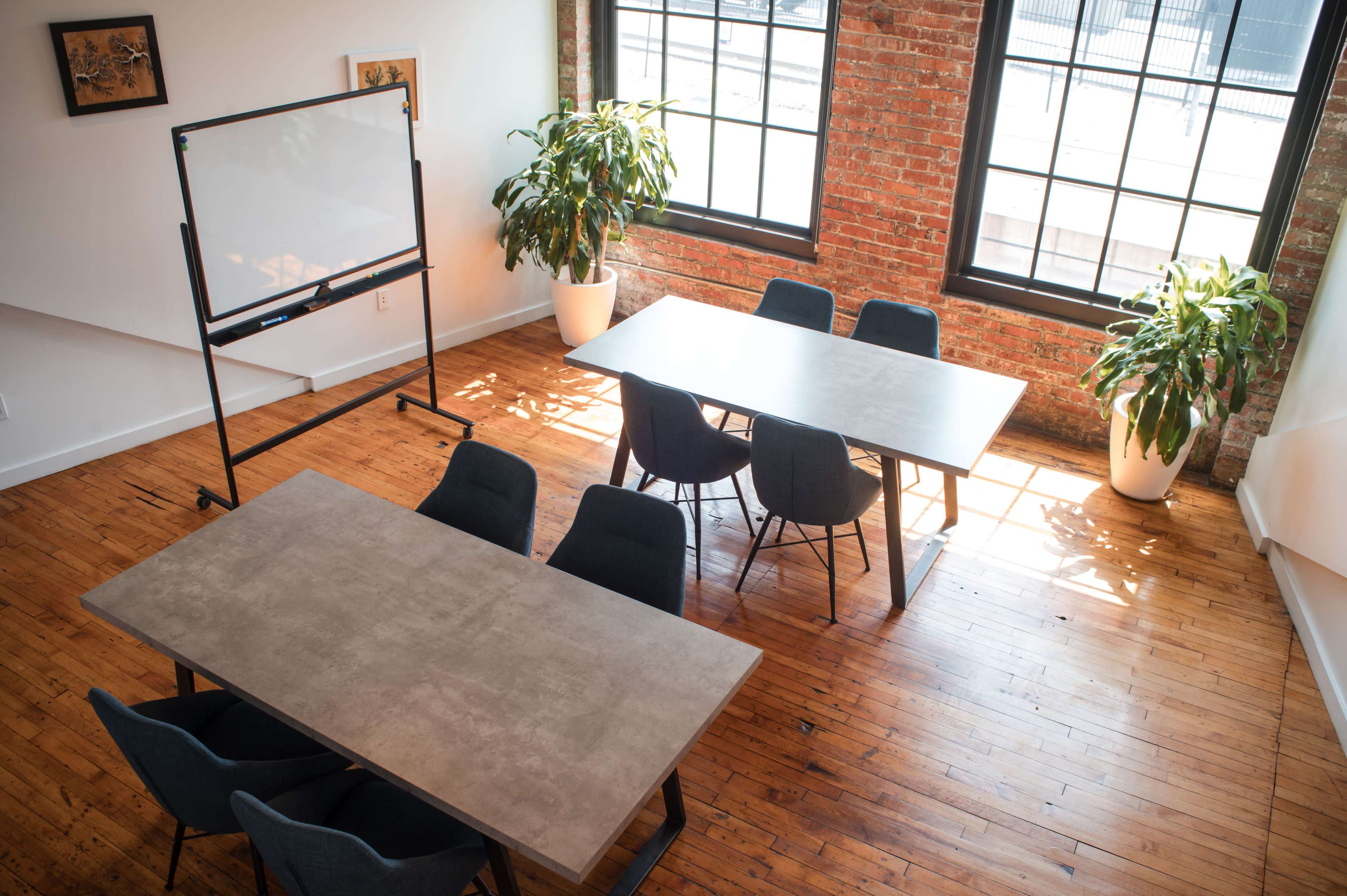 A bright meeting room features two large tables surrounded by chairs, with green plants and large windows letting in natural light.