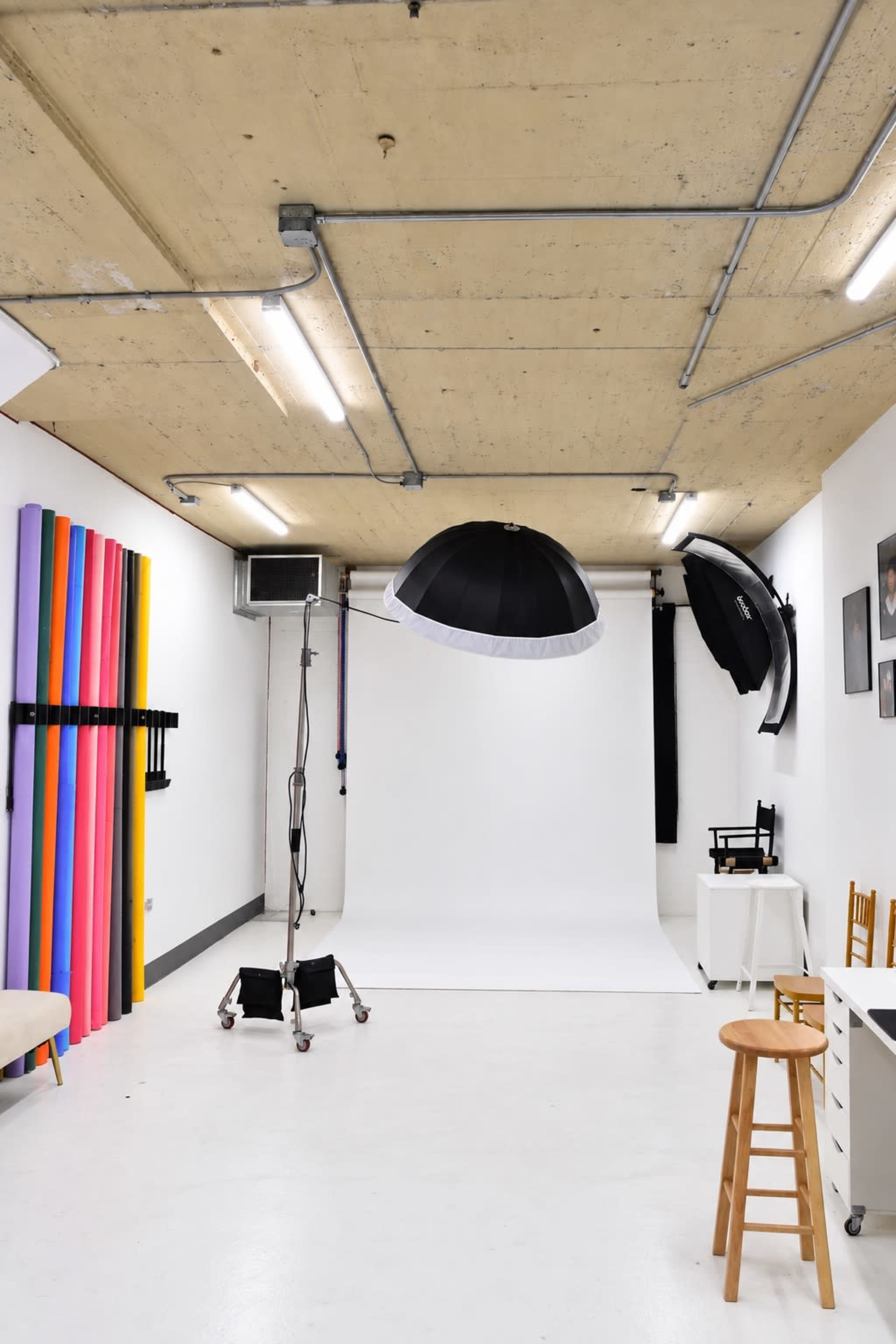 The image shows a photography studio with a white backdrop, lighting equipment suspended from the ceiling, and colorful backdrop rolls on one wall.