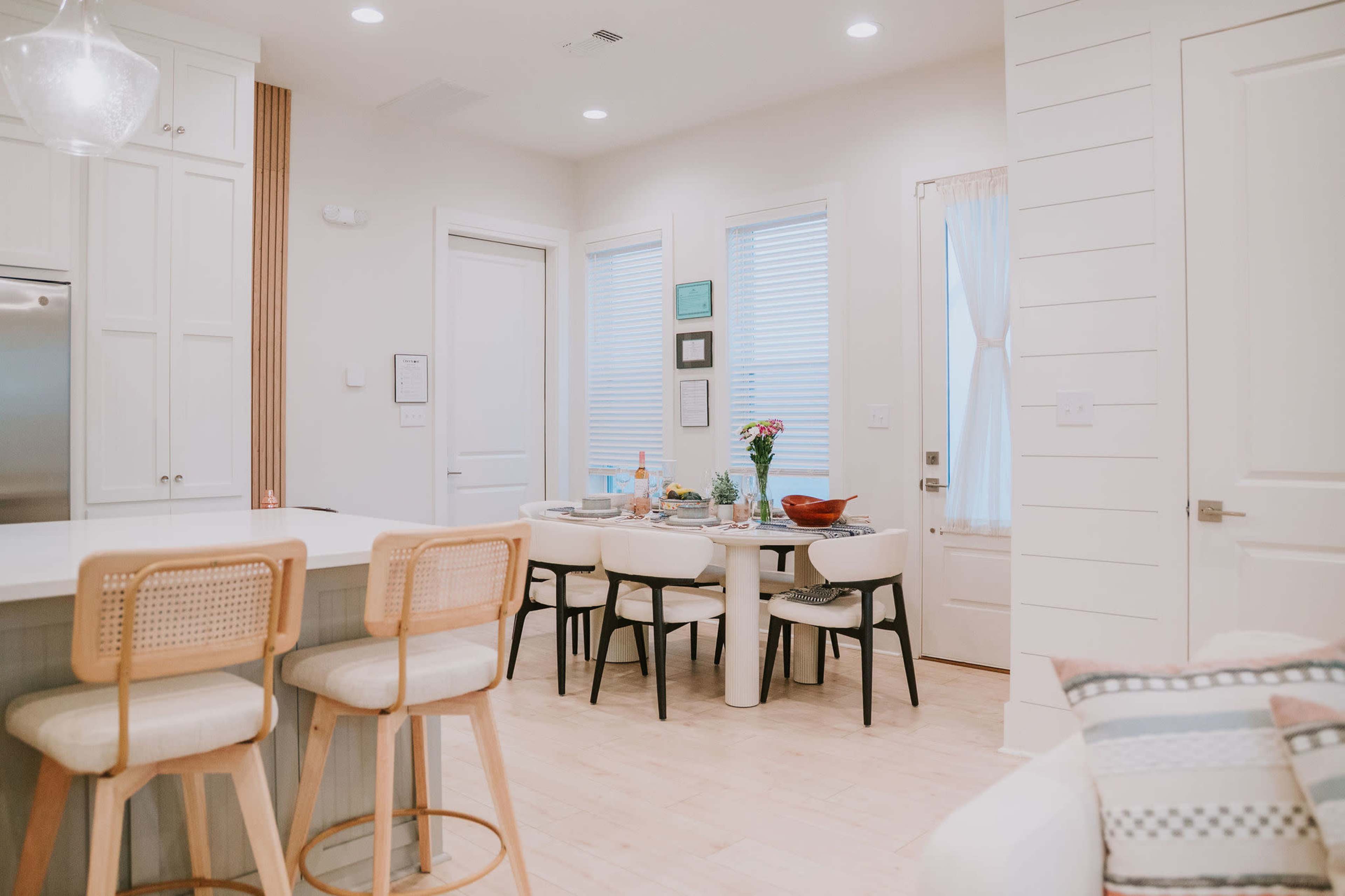 A modern kitchen and dining area features a table set for a meal, surrounded by six chairs, with bar stools at a kitchen island nearby.