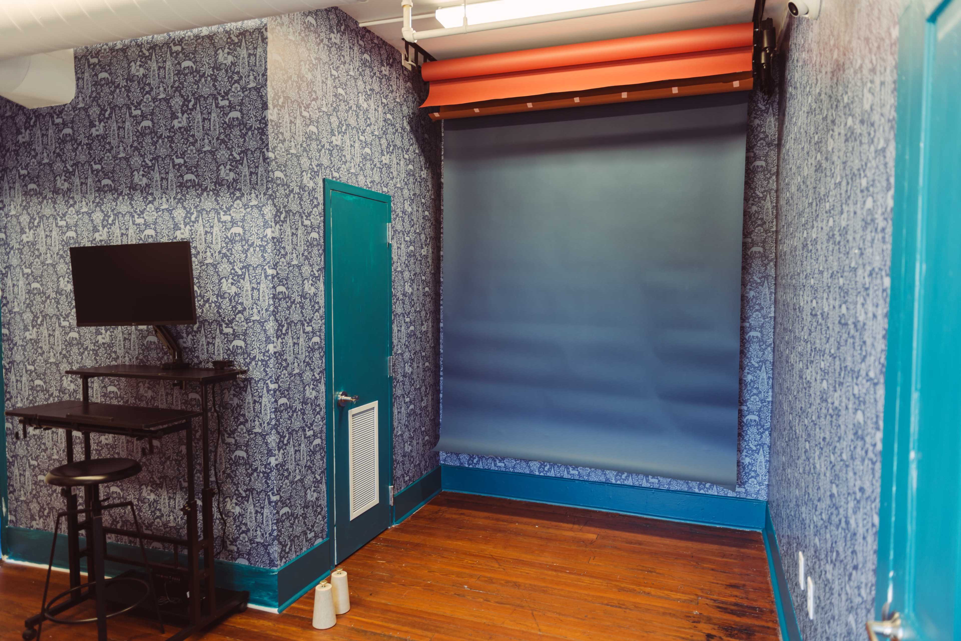 A small room with patterned blue wallpaper, a blue backdrop, a wall-mounted monitor, and a wooden floor.