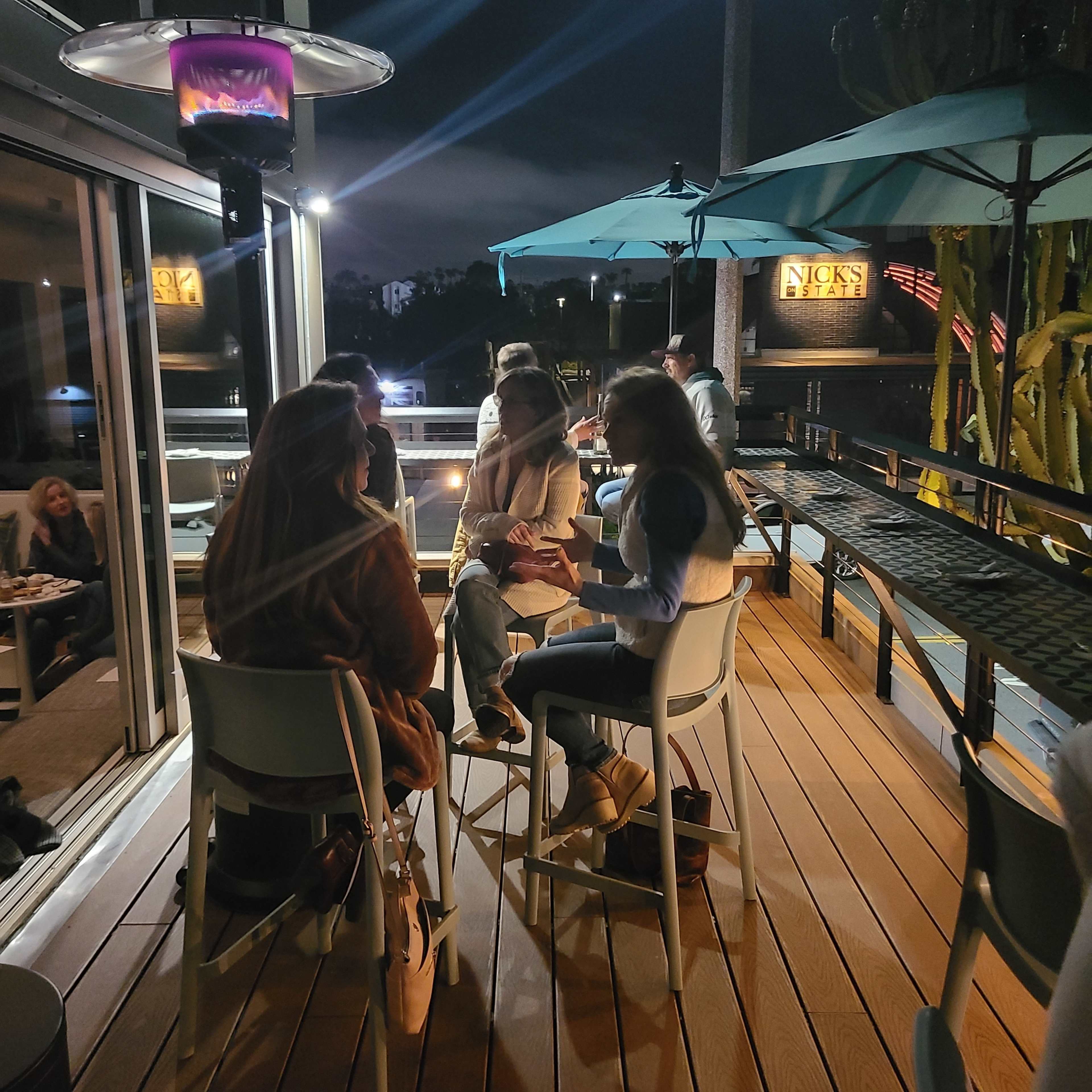 A group of women sits around a table on a balcony, chatting while illuminated by a heating lamp at night.