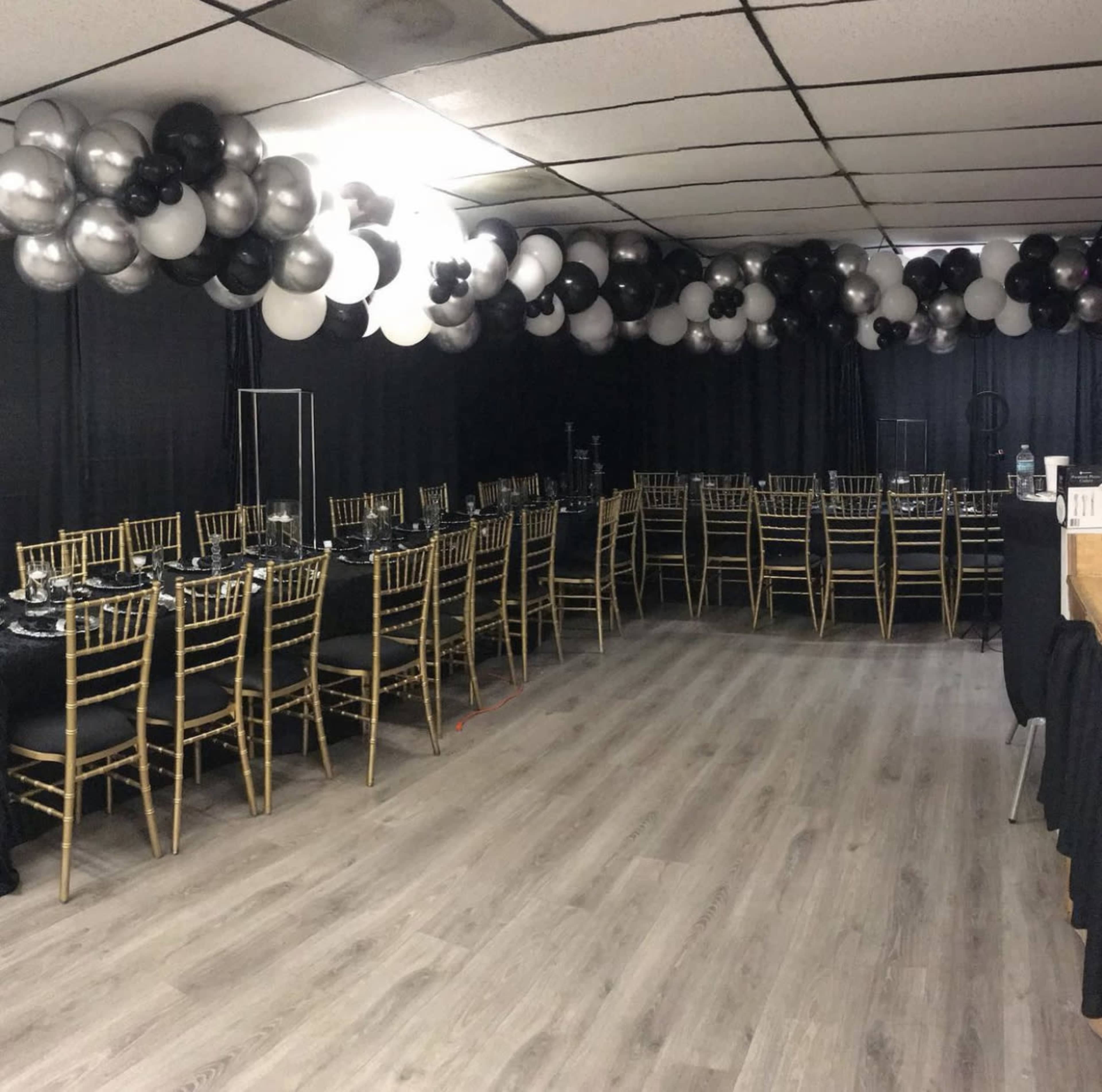 A dining area is set up with multiple rows of gold Chiavari chairs and black tables, decorated with silver and black balloons overhead.