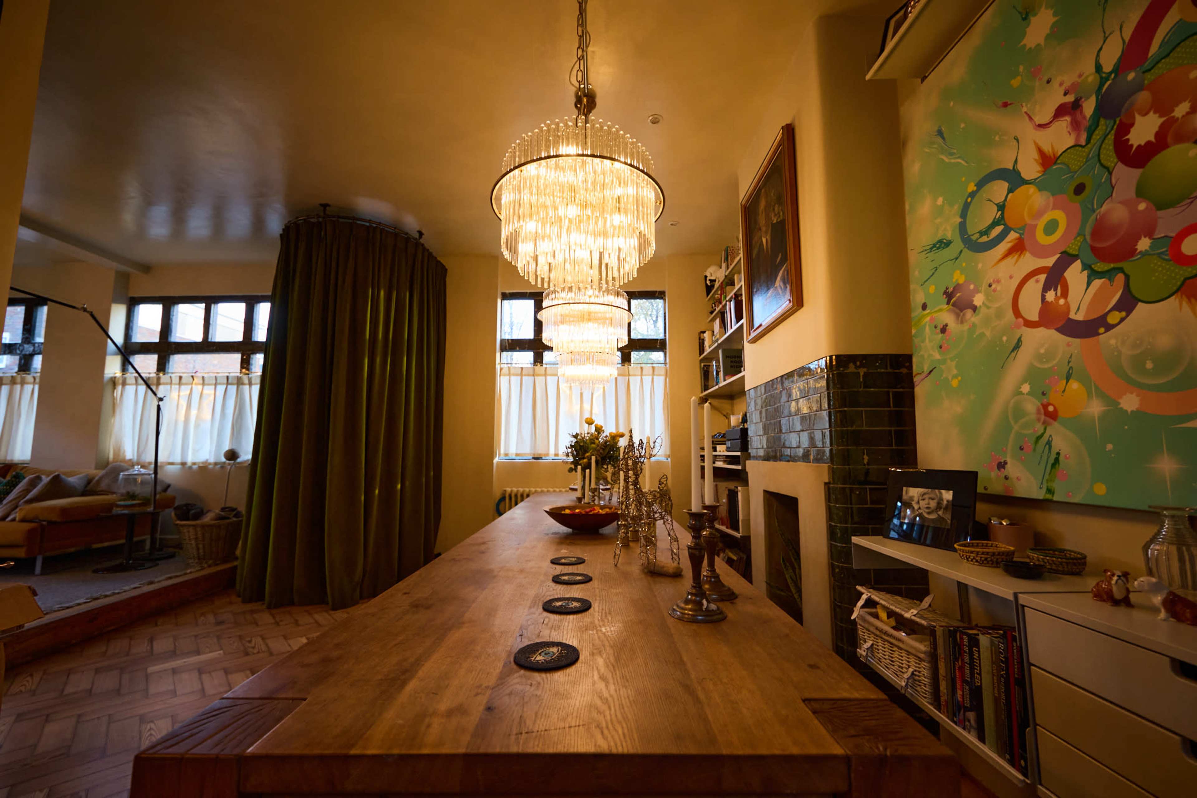 The image shows a spacious dining area featuring a long wooden table, elegant chandeliers, and colorful artwork on the wall.