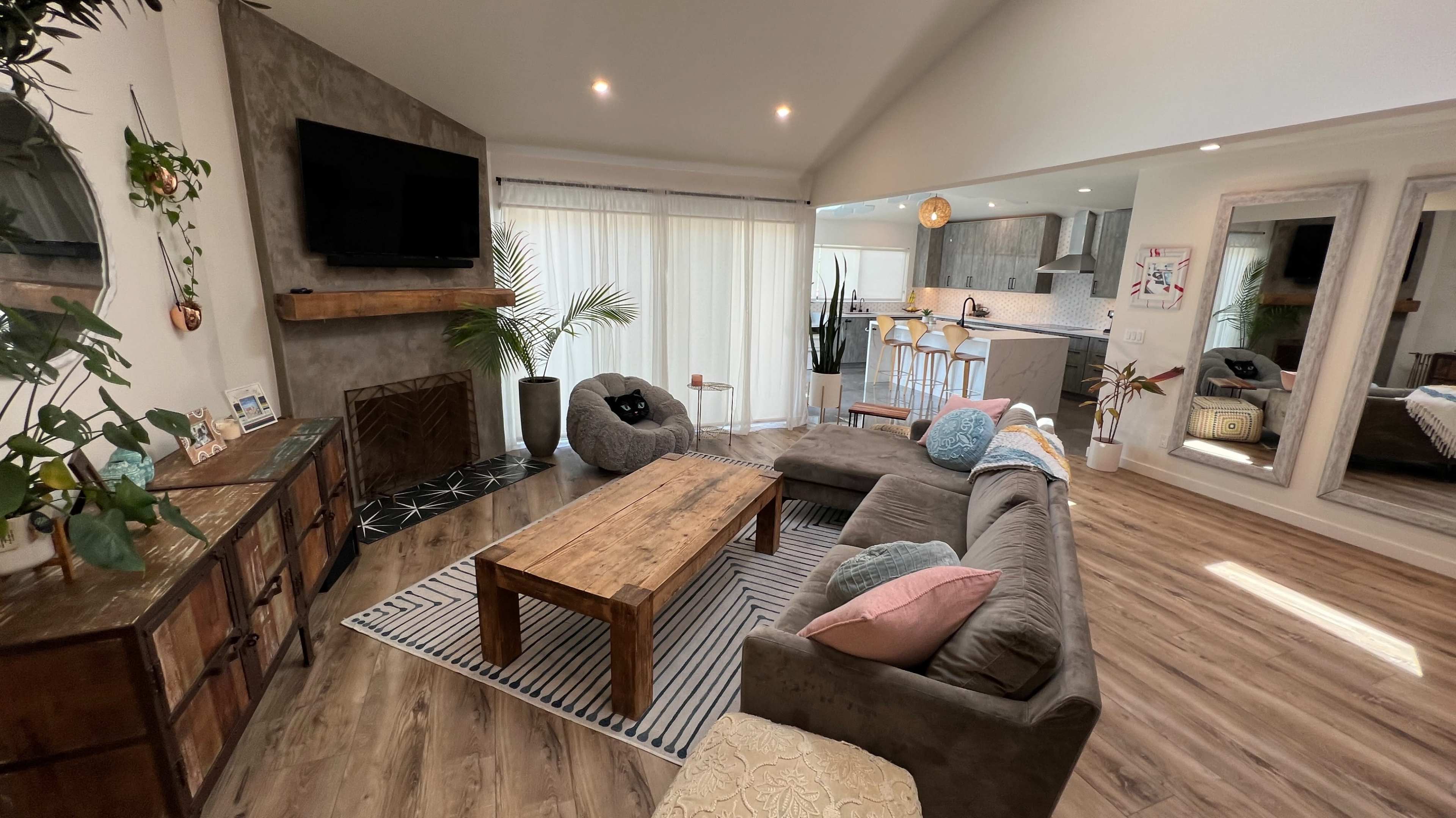 A modern living room with a sectional sofa, a wooden coffee table, a television mounted on the wall, and large windows displaying natural light.