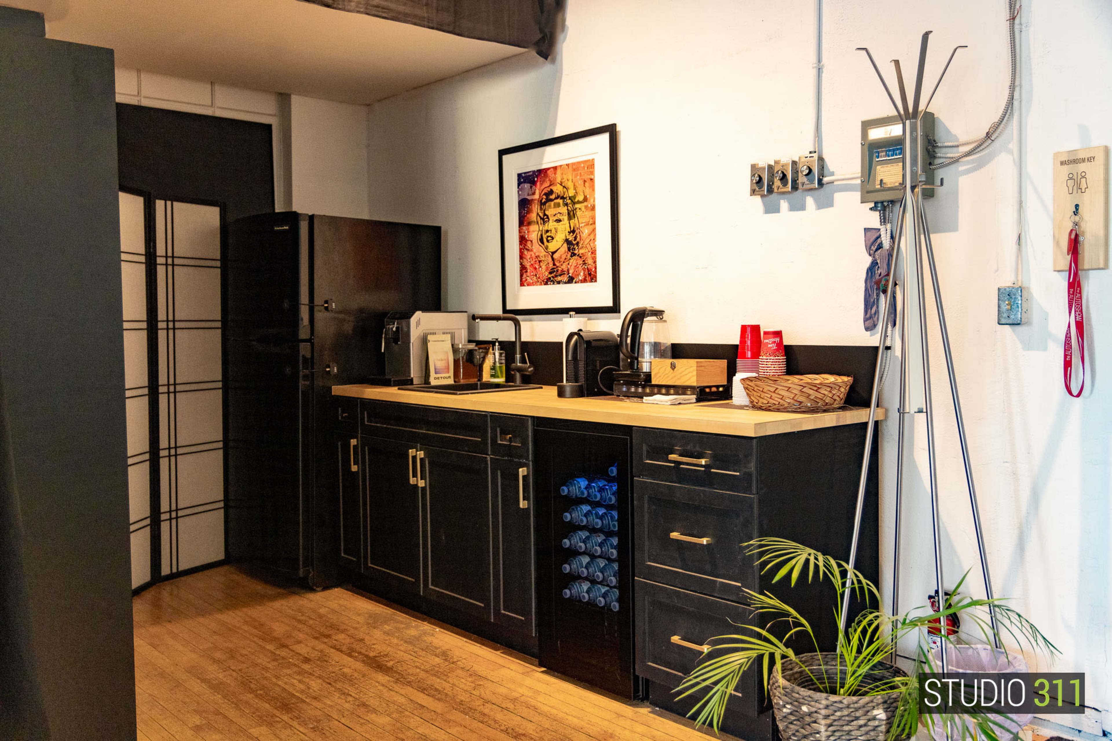 The image shows a compact kitchen area with dark cabinets, a countertop featuring a coffee maker, and a refrigerator, along with a wall-mounted shelf and a plant in the corner.