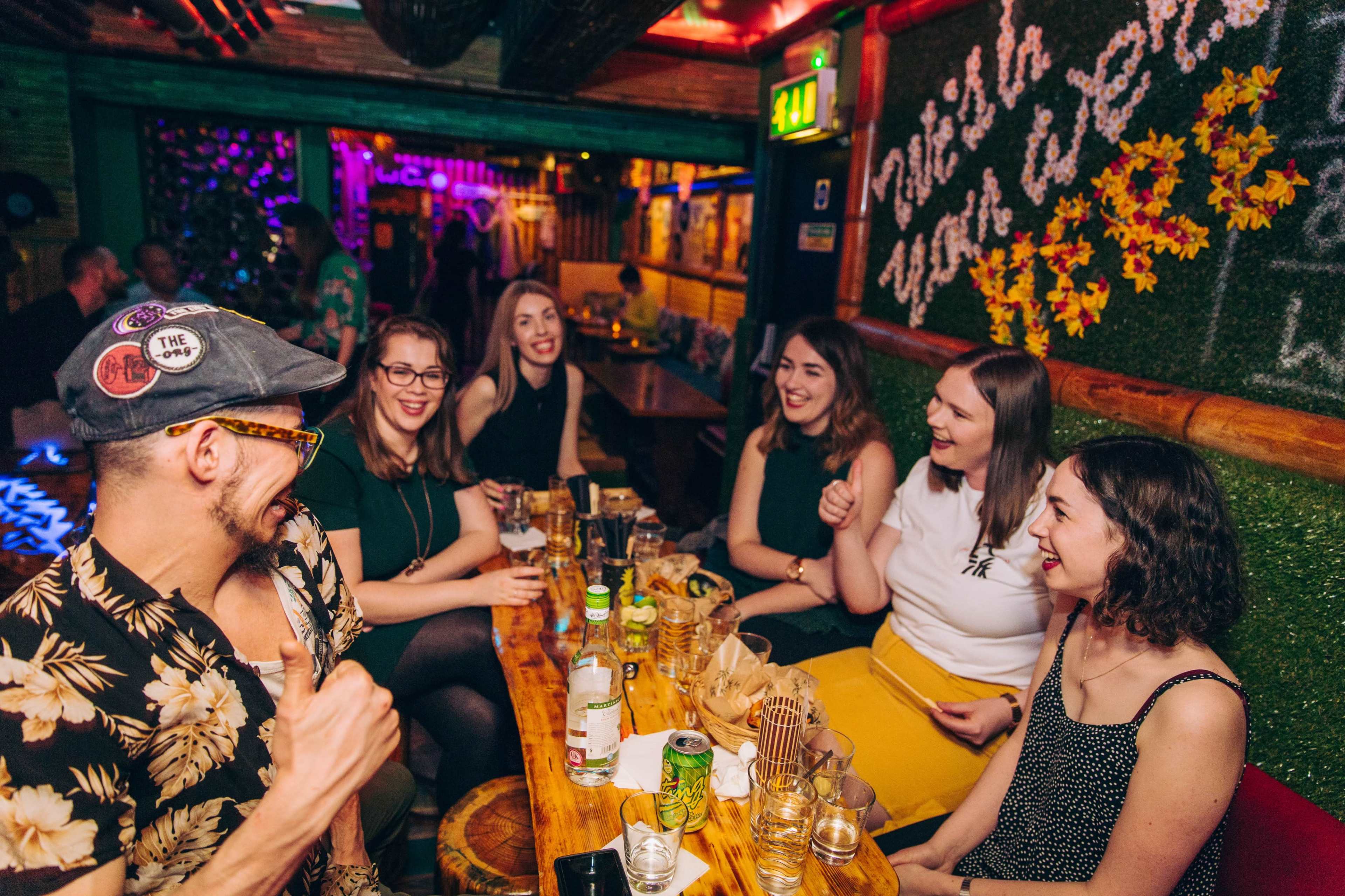 A group of six people sits at a table in a lively bar, engaging in conversation and surrounded by colorful decorations.