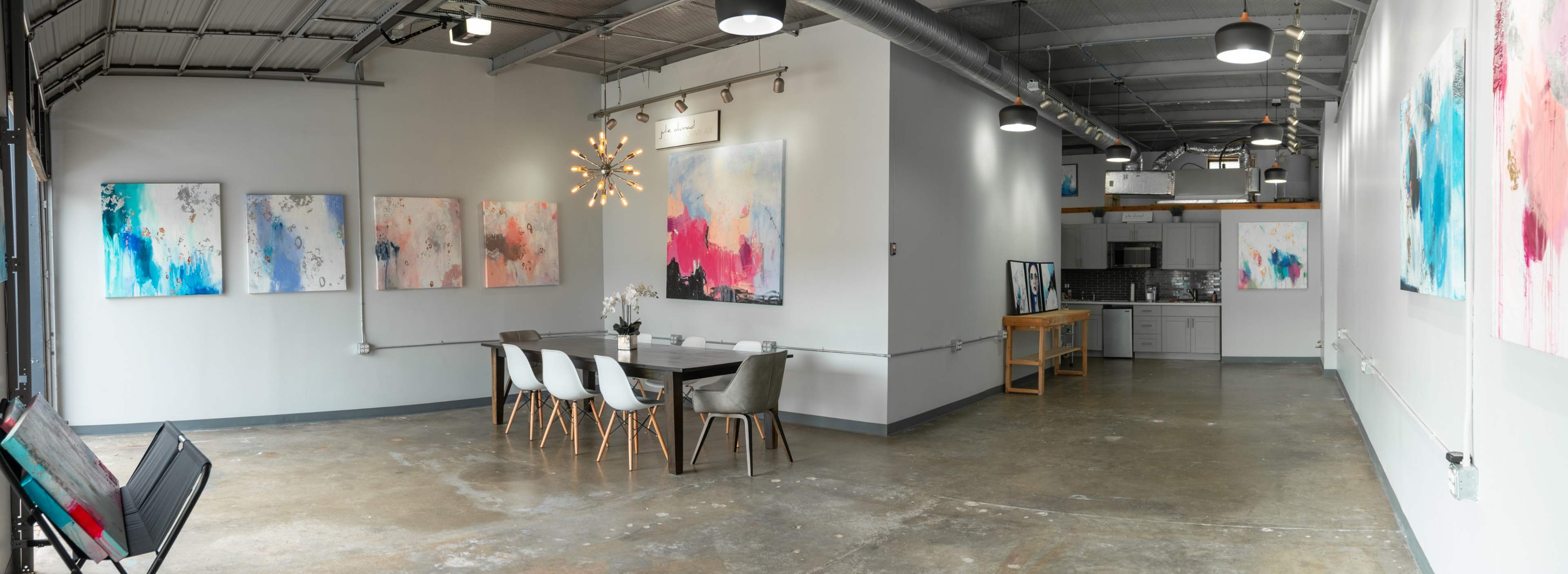 A modern studio space with abstract paintings on the walls, a dining table with chairs in the foreground, and a kitchenette in the background.