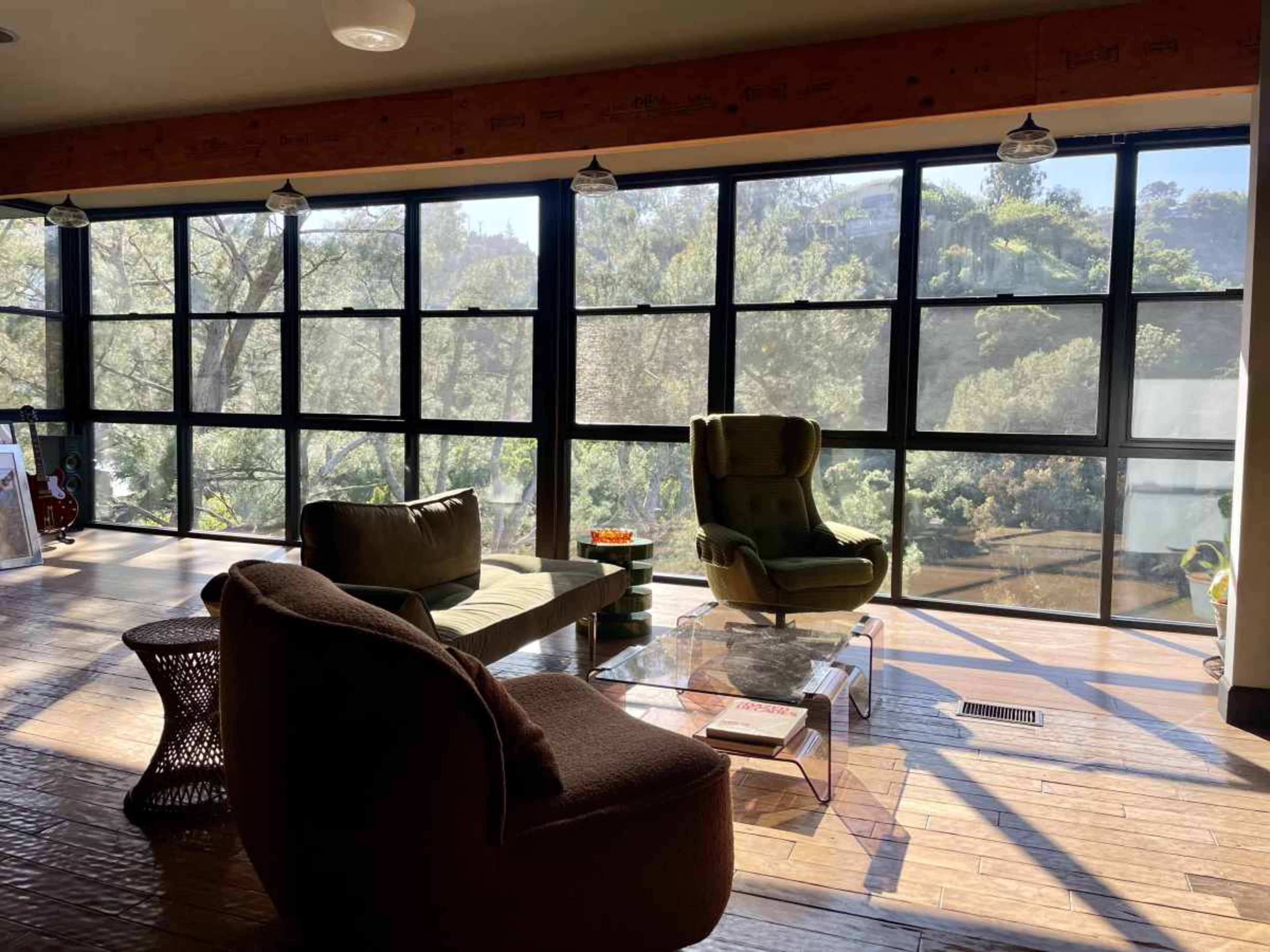 The image shows a cozy sitting area with two armchairs and a sofa beside large windows that offer a view of greenery outside.