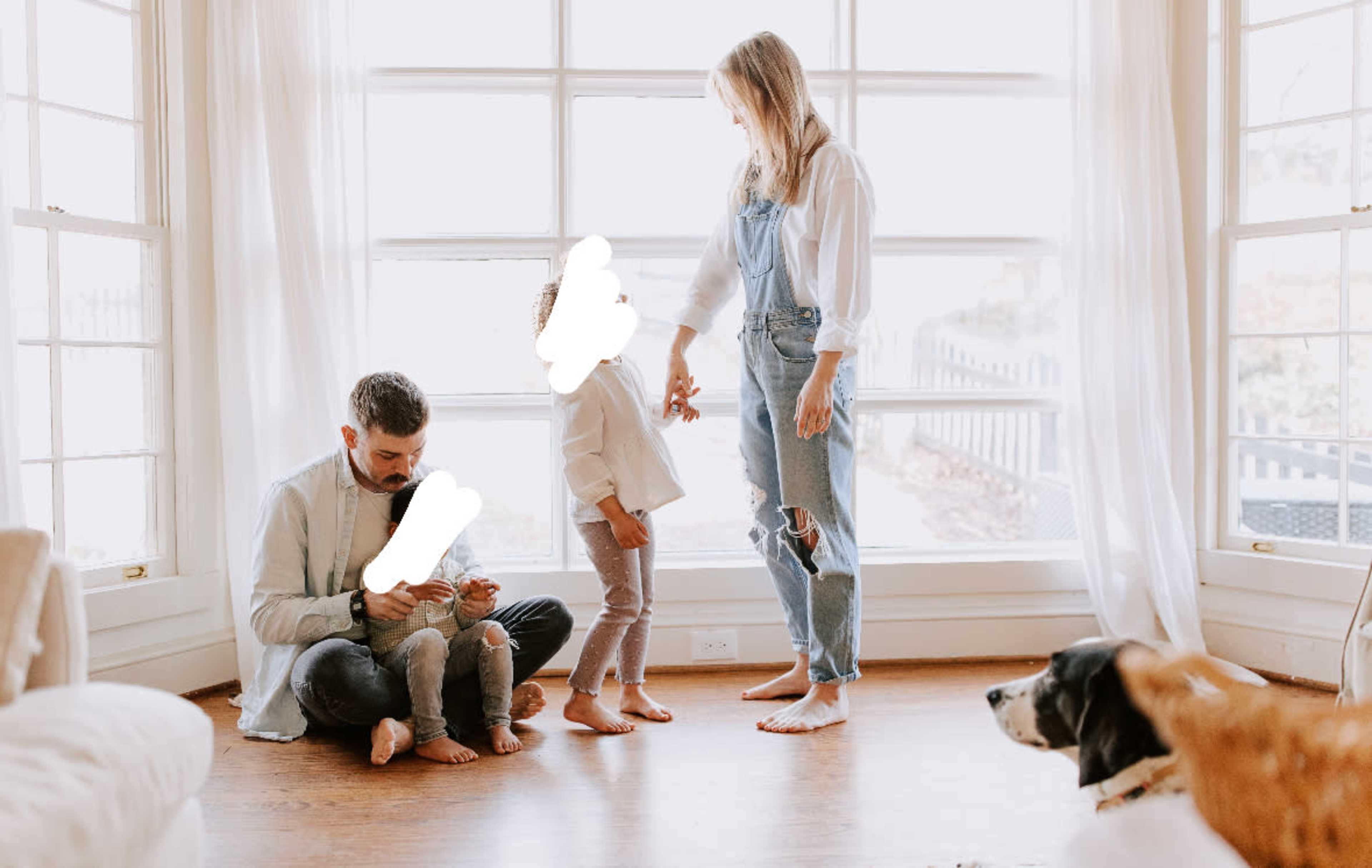 A family sits in a sunlit living room, with one parent on the floor using a phone, a child standing beside them, and the other parent interacting while a dog watches.
