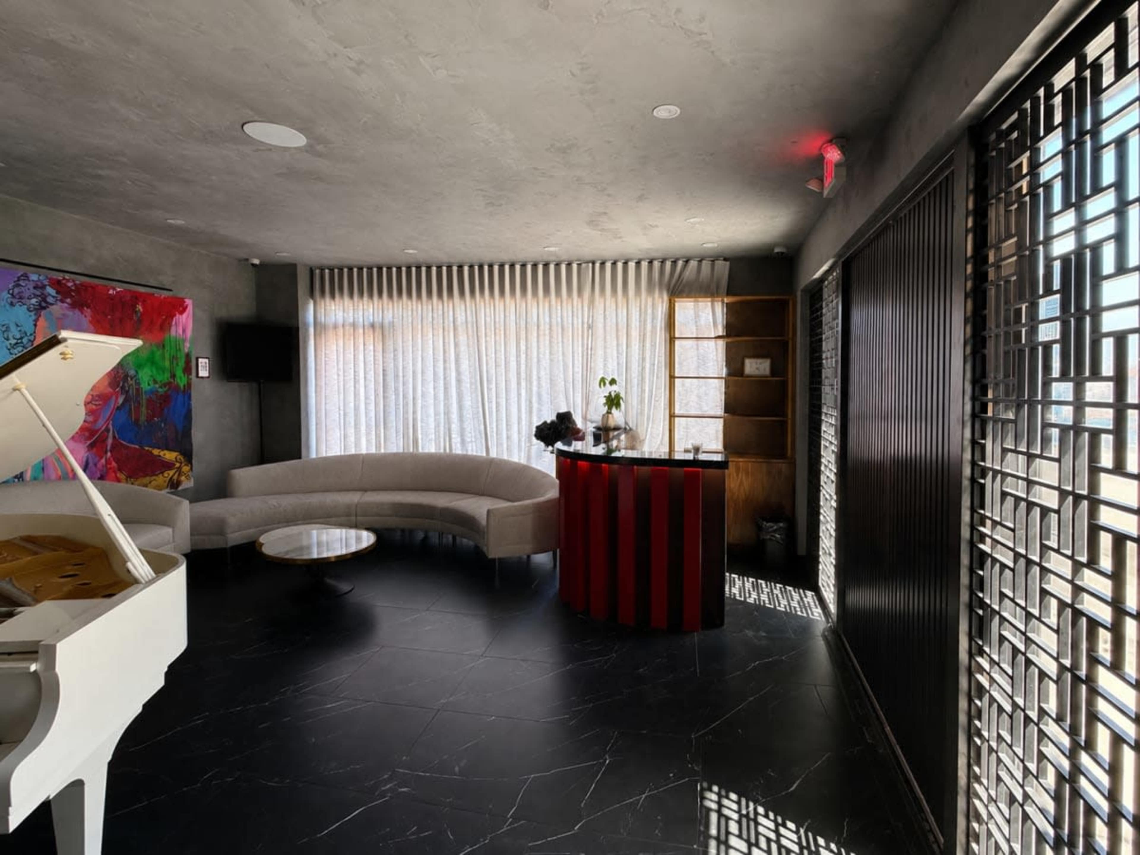 The image shows a modern interior space featuring a curved sofa, a small round table, a white piano, and a reception desk with red vertical stripes, all illuminated by natural light filtering through sheer curtains.