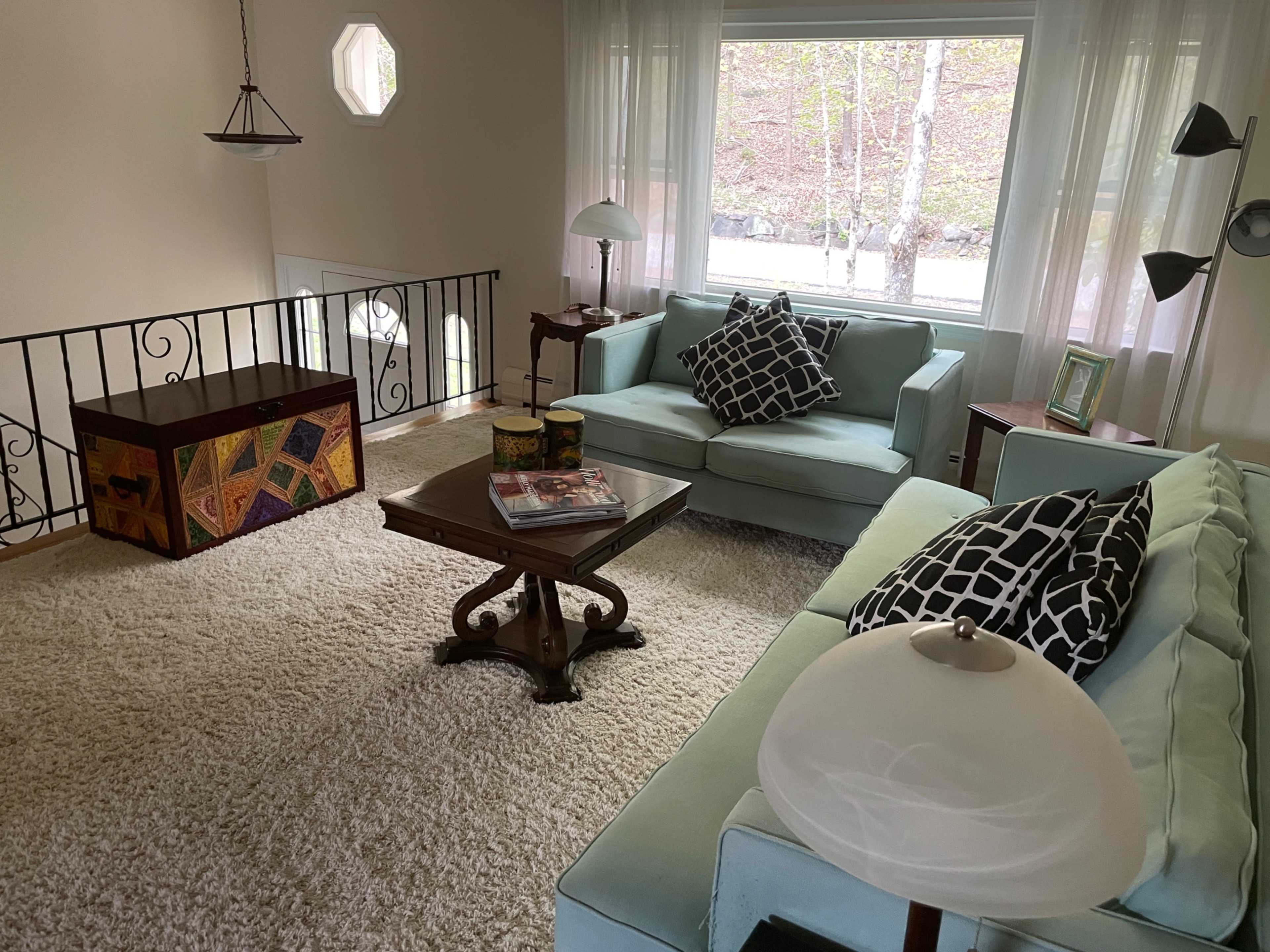 A cozy living room features light blue furniture, a patterned rug, and a large window overlooking a tree-lined view.