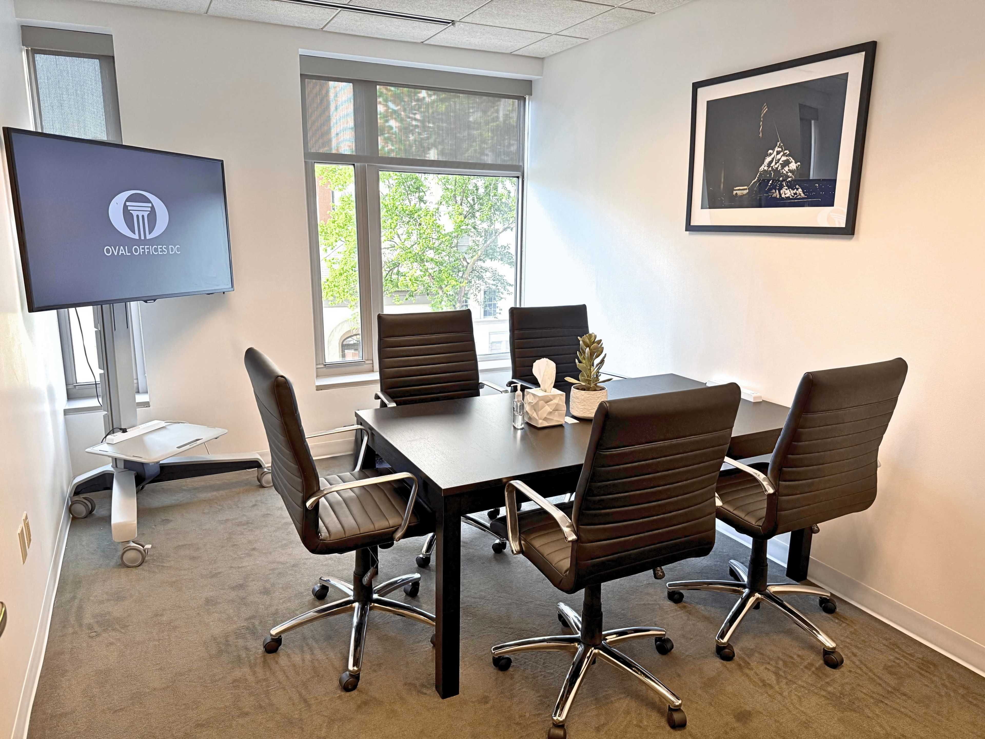 The image shows a small conference room with a table surrounded by five black office chairs, a monitor on a stand, and large windows allowing natural light.