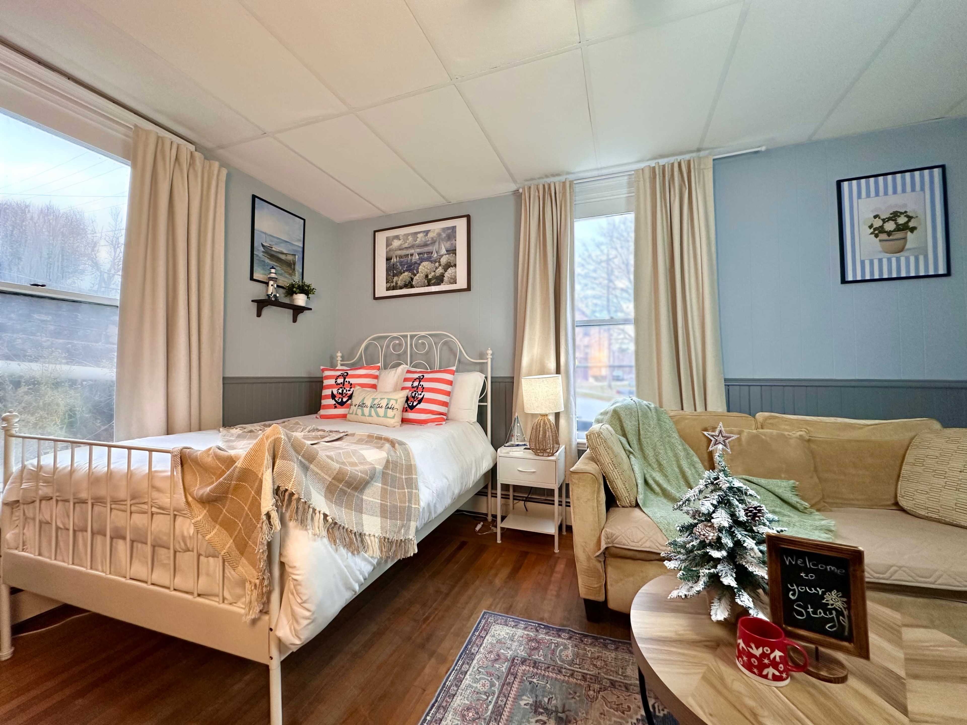 The image shows a small, cozy bedroom with a white metal bed, a beige couch, and a small decorative Christmas tree on a round table.