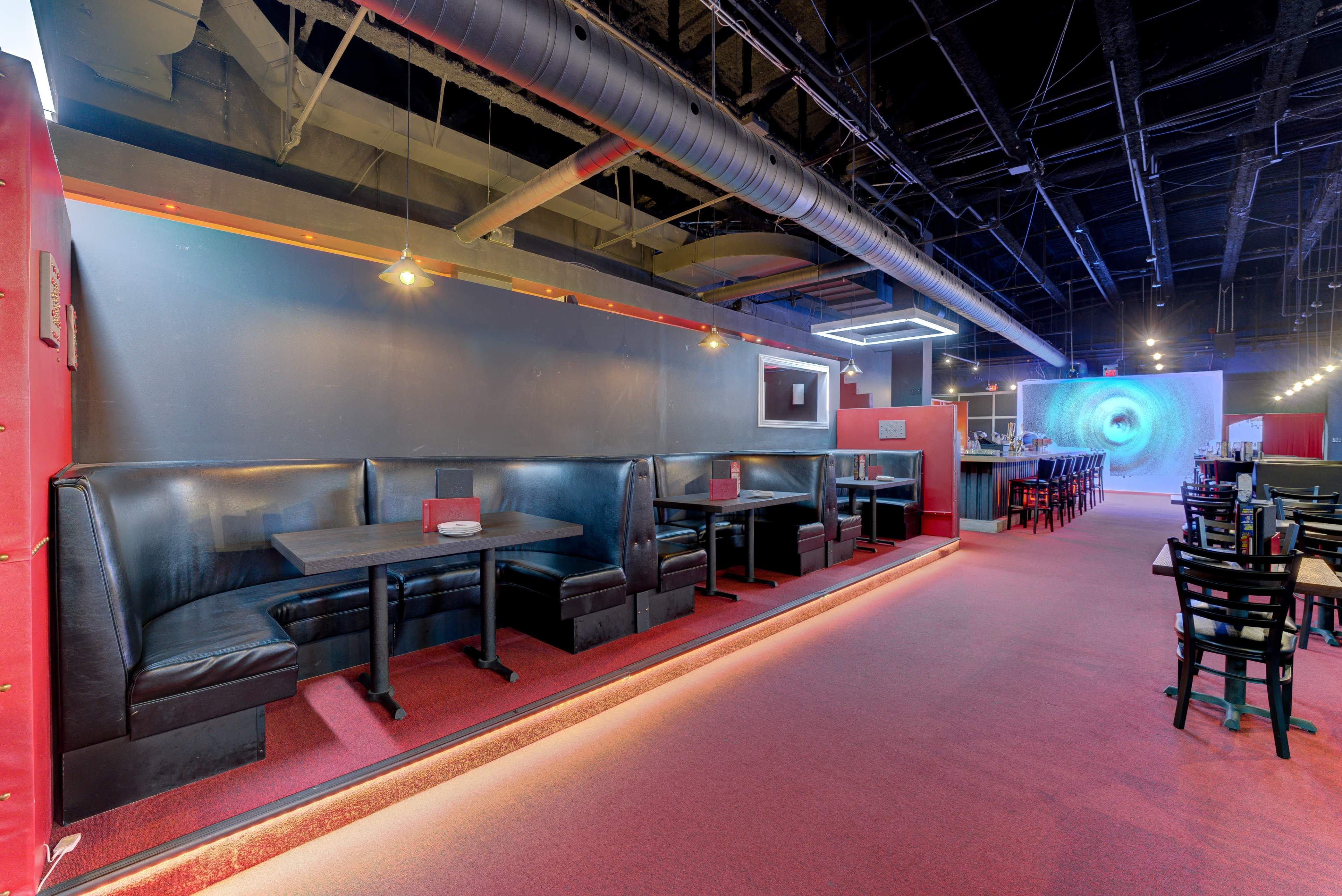 A modern restaurant interior with black booths and red carpeting, leading to a bar area and a colorful wall decoration in the background.