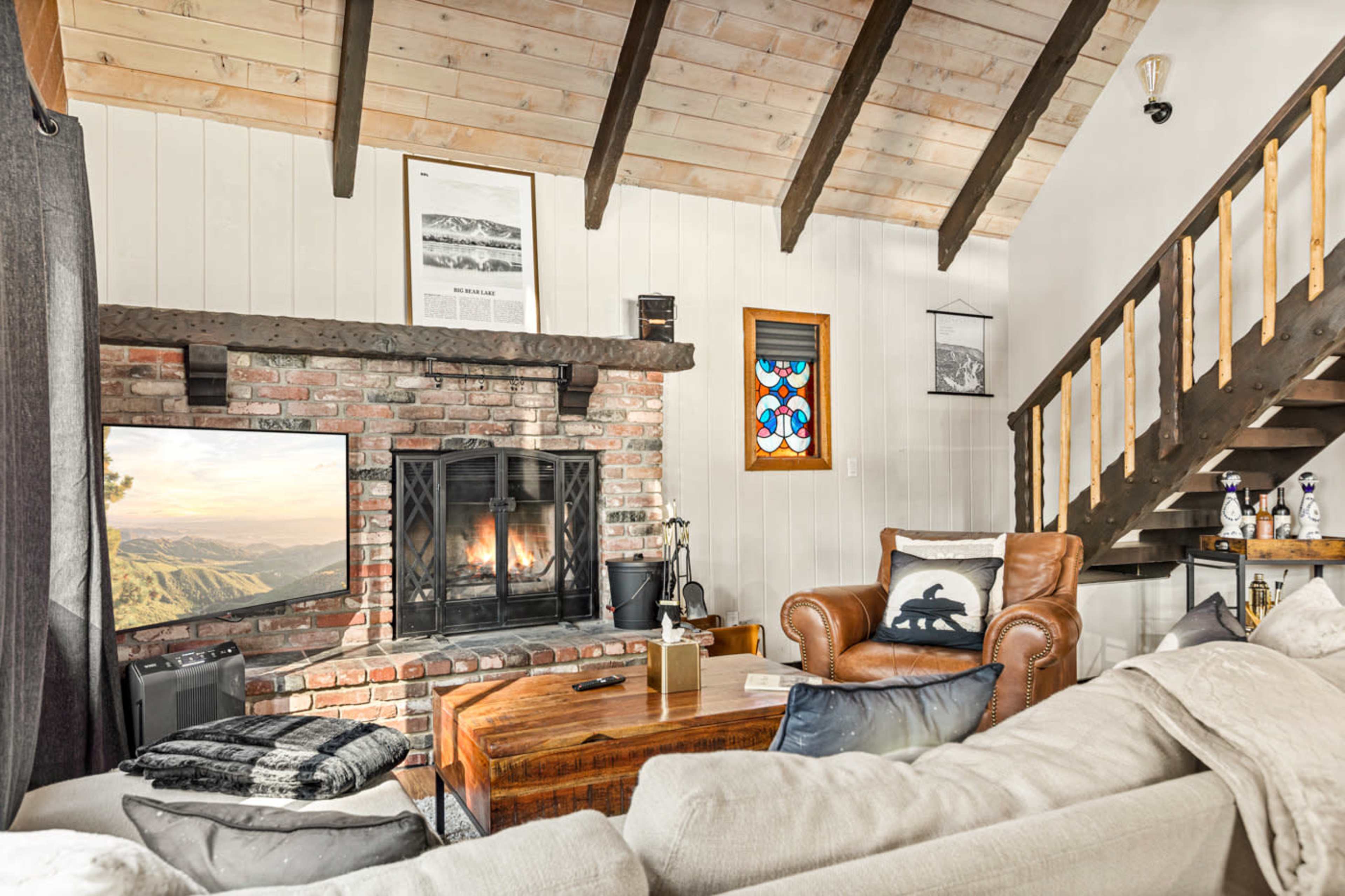 A living room features a brick fireplace with a television, a leather armchair, a wooden coffee table, and a staircase leading to the upper level.