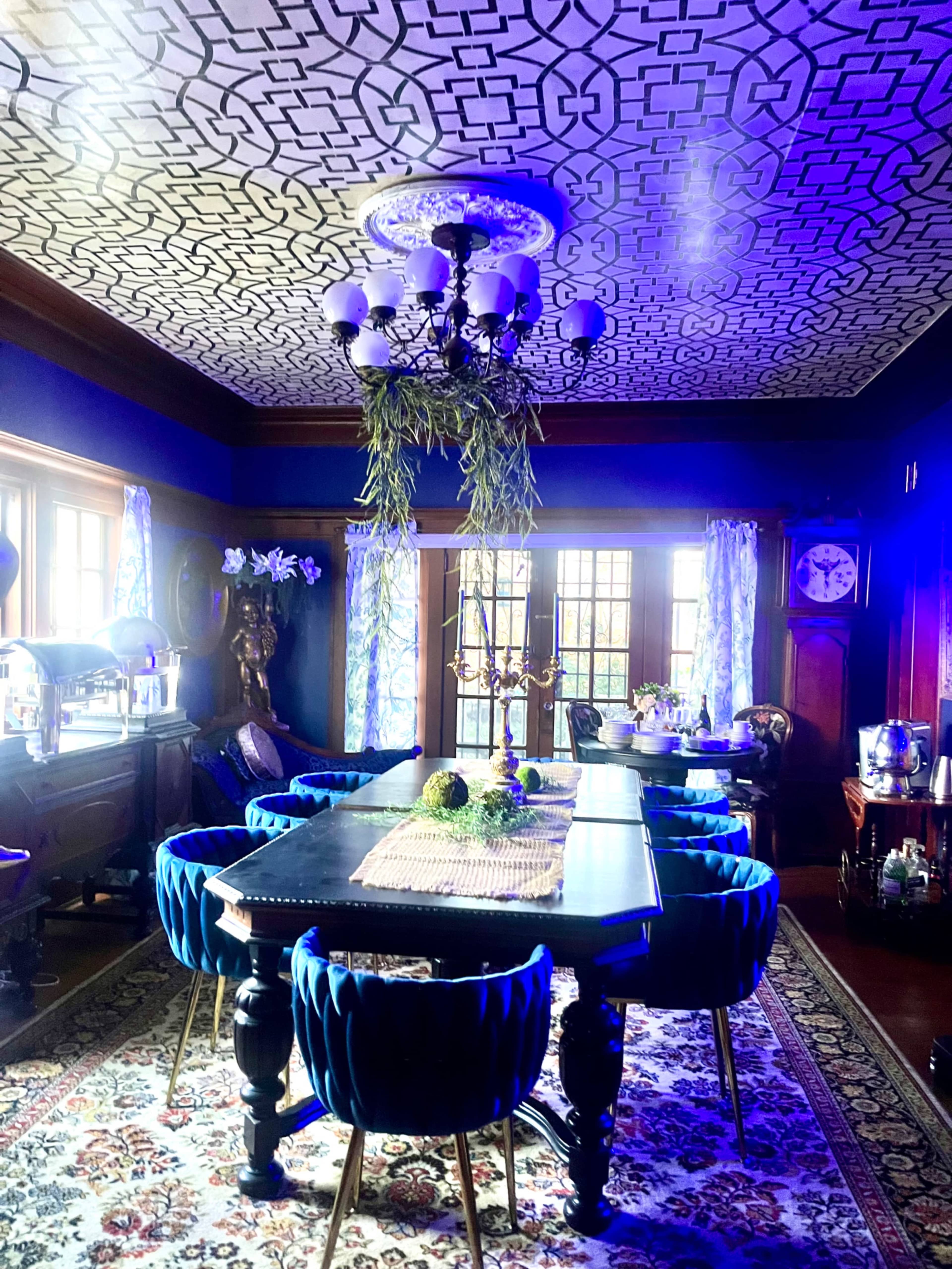 The image shows a spacious dining room featuring a large black table surrounded by blue velvet chairs, with a patterned ceiling and a chandelier above.