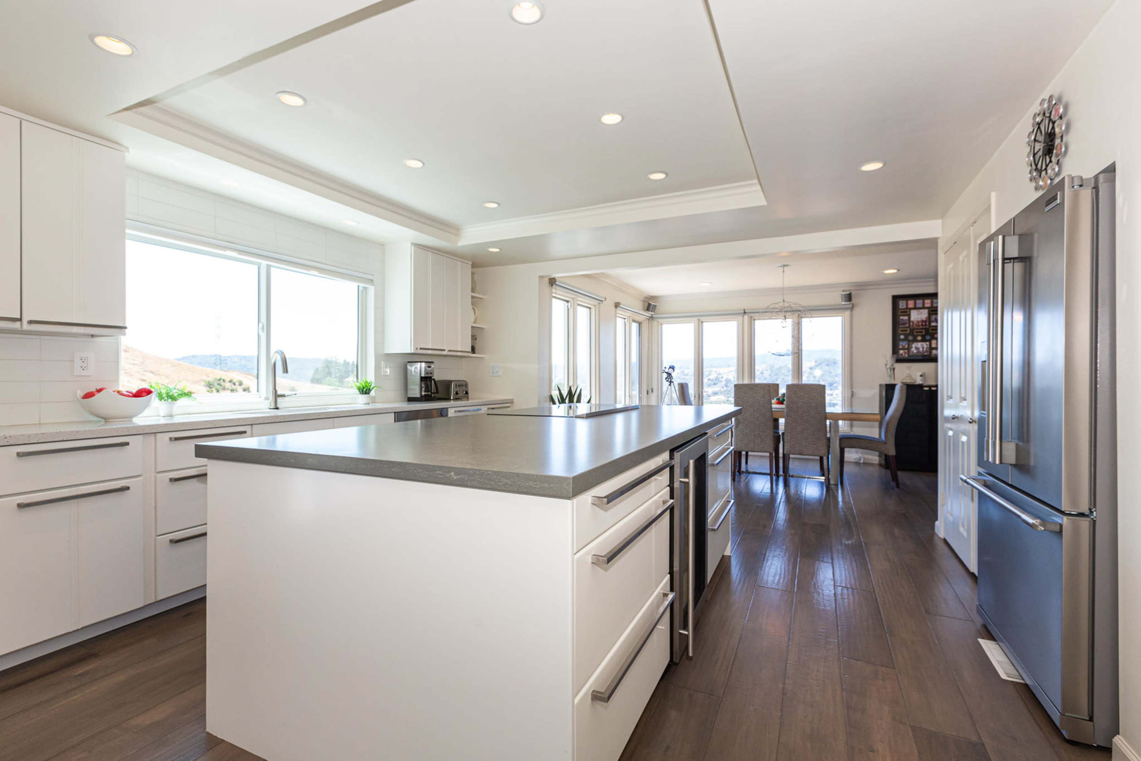 A modern kitchen with a central island, featuring white cabinetry, stainless steel appliances, and large windows overlooking a landscape.