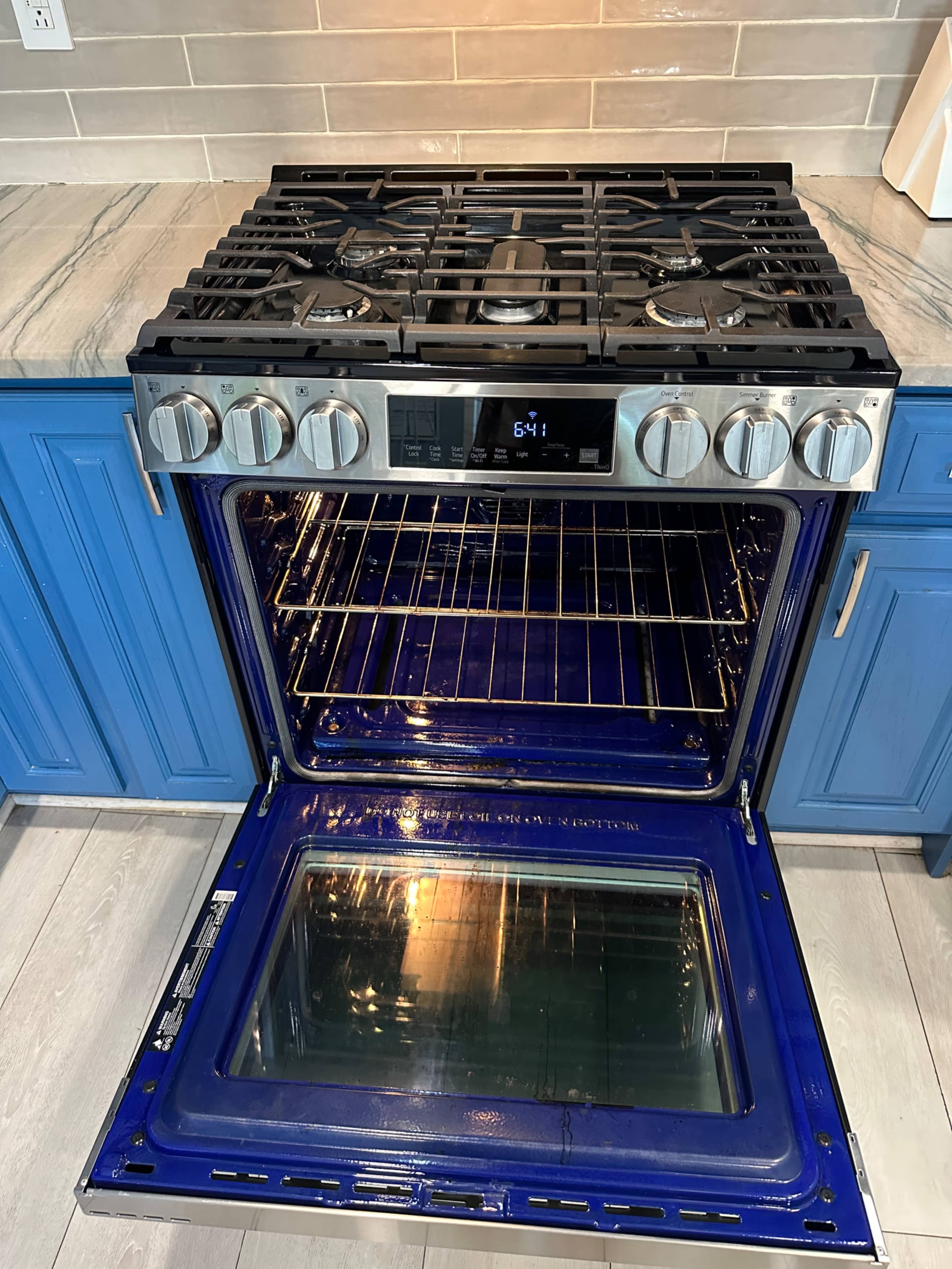 The image shows a gas range oven with a blue interior, including a visible oven door that is open, revealing empty racks inside.
