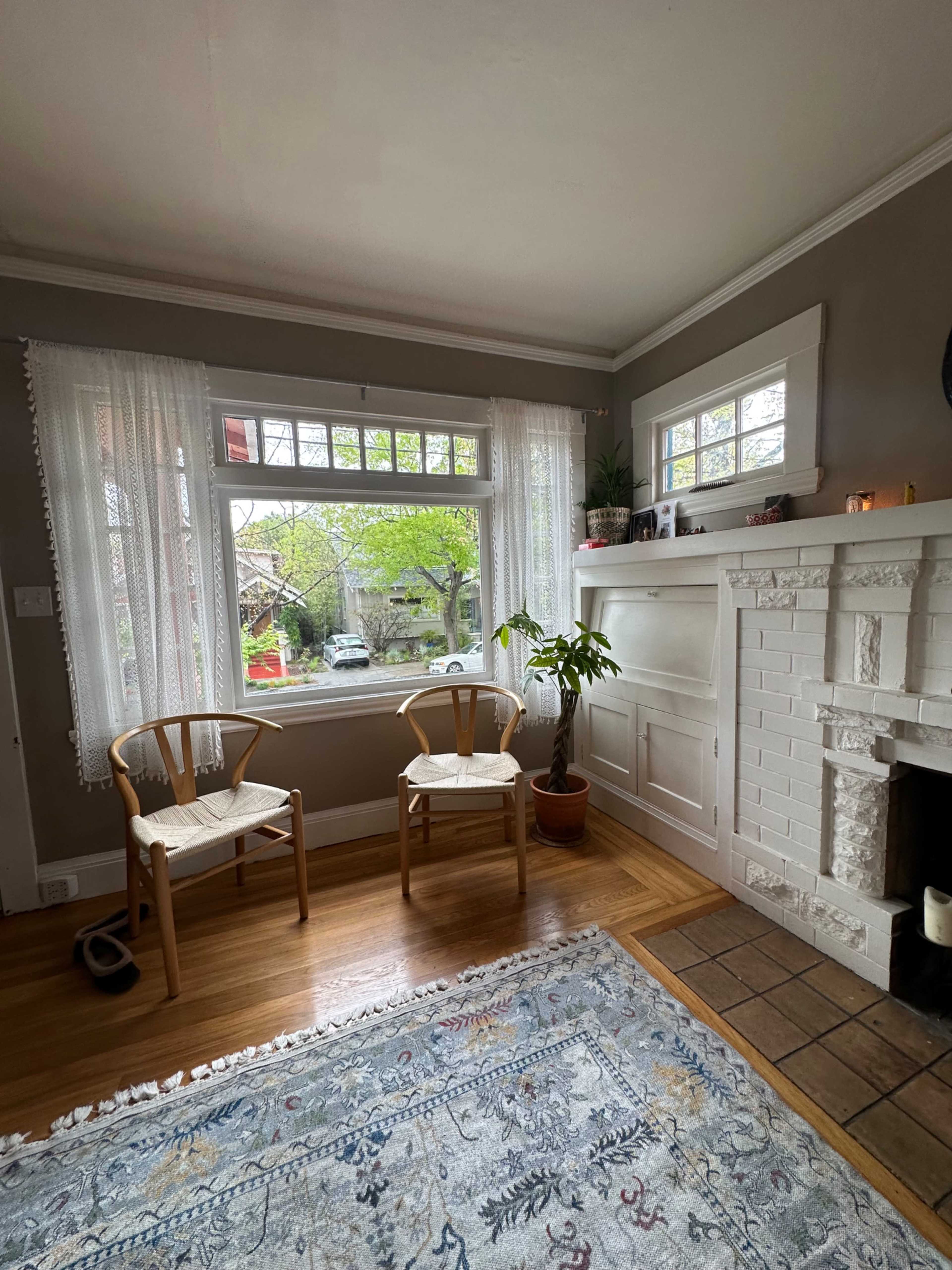 The image shows a living room with two wooden chairs near a window, a potted plant beside them, and a white brick fireplace.