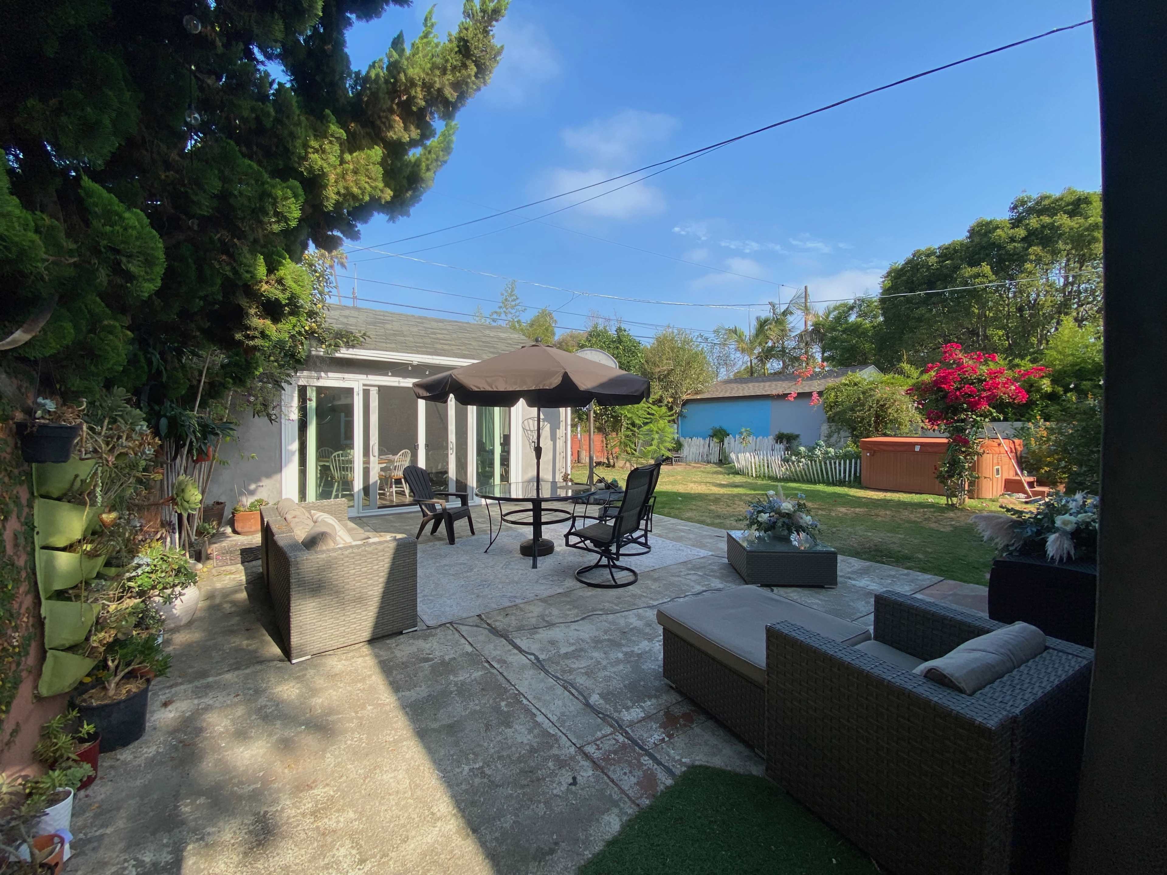 A backyard patio area with outdoor furniture, a table and umbrella, and a grassy lawn surrounded by trees and colorful plants.