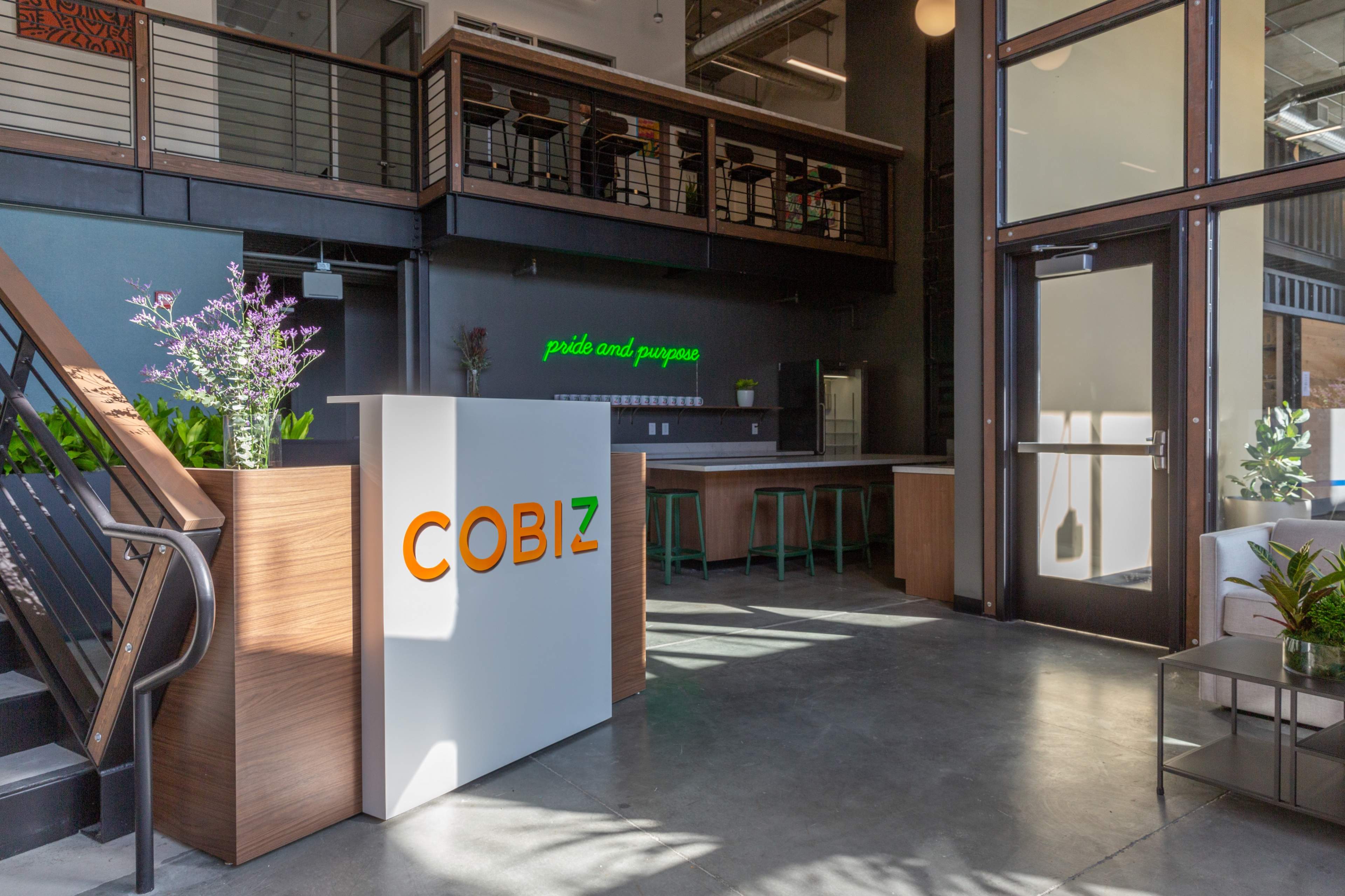 A modern office reception area featuring a white front desk with the word "COBIZ" in orange and green, along with indoor plants and a seating area.