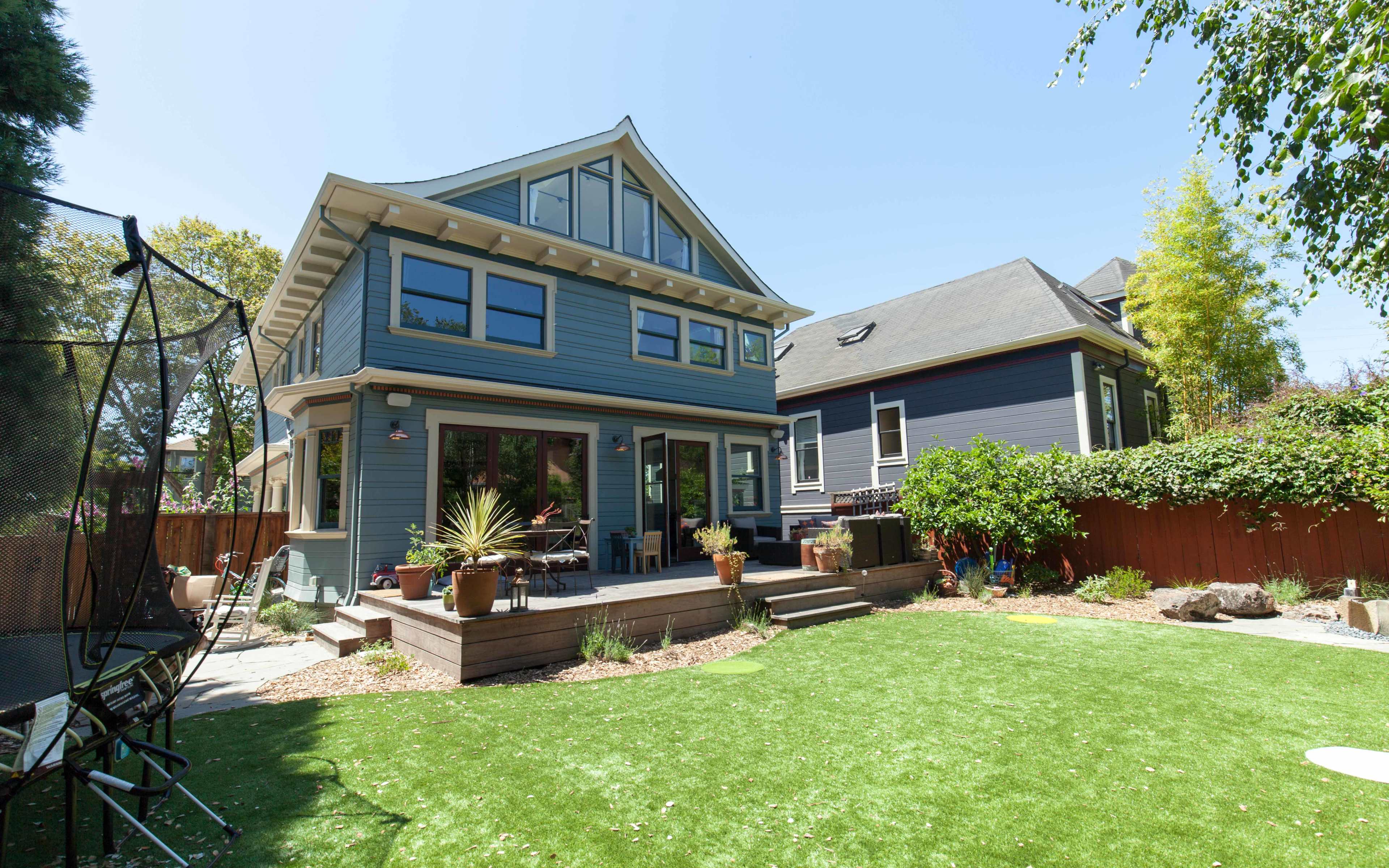 A two-story blue house with large windows and a patio overlooks a well-maintained backyard with artificial grass and various plants.