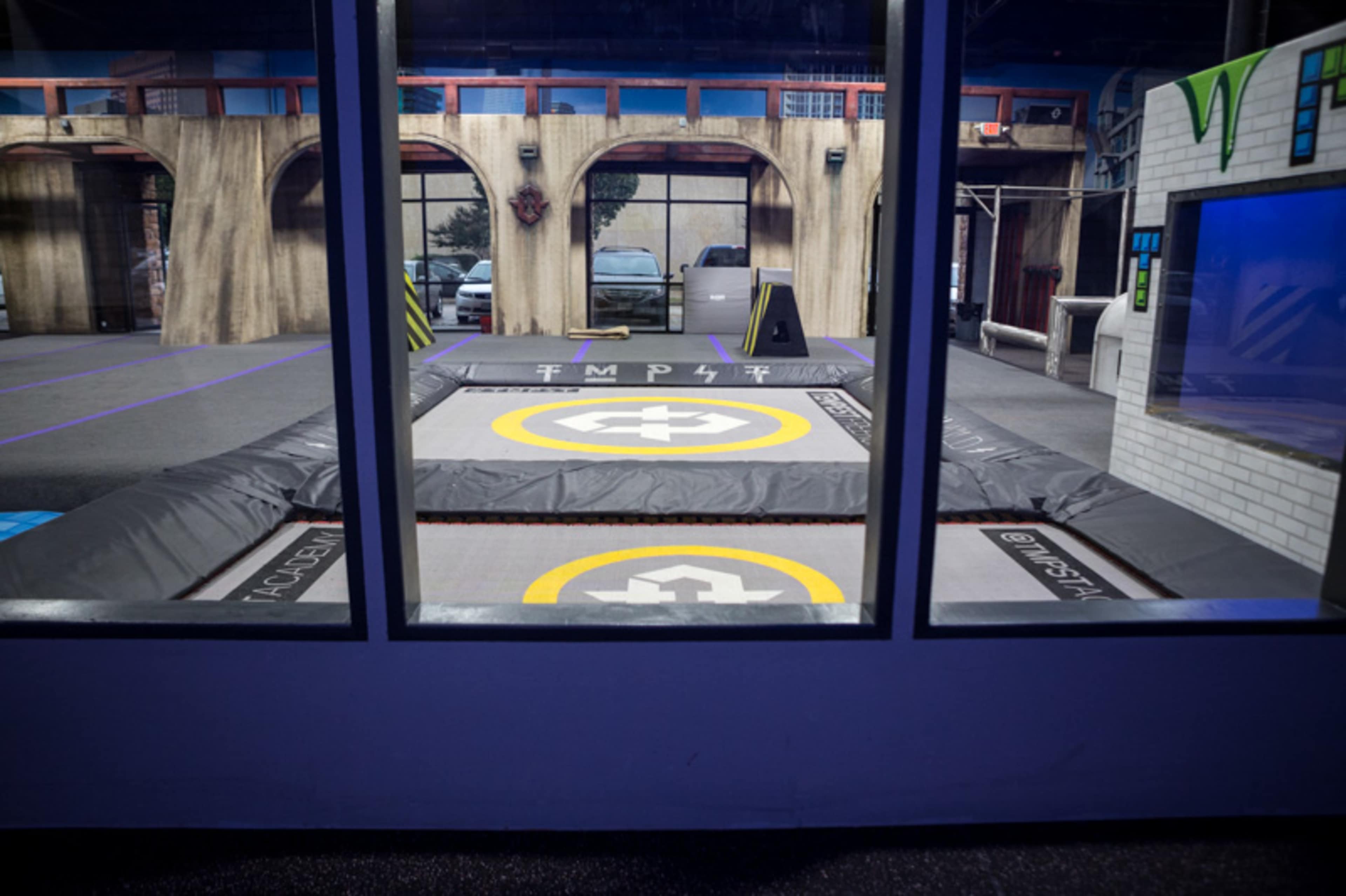 The image shows an indoor trampoline area with multiple trampolines and a surrounding urban-themed environment visible through large windows.