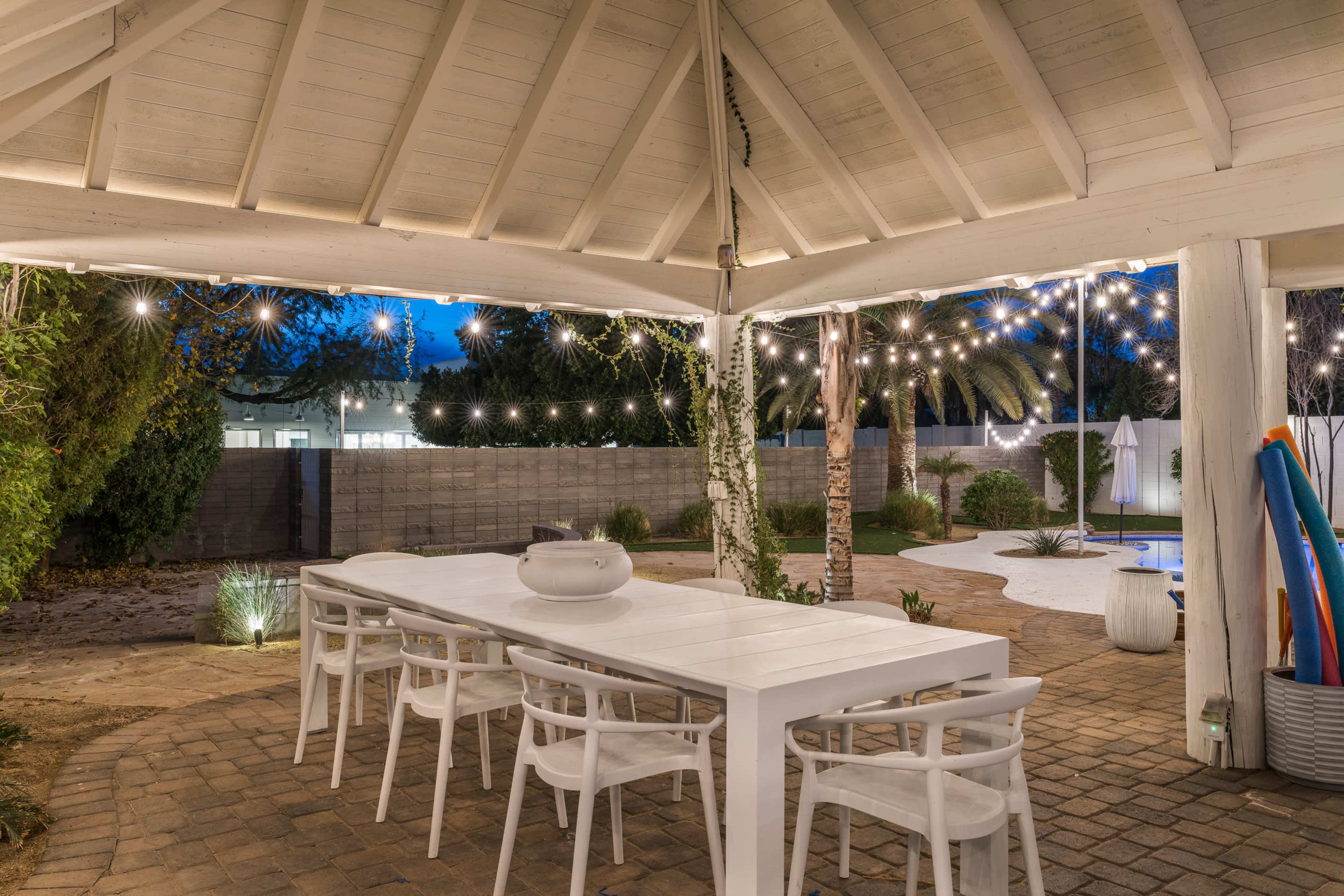 A modern outdoor dining area with white furniture, string lights, and a view of a landscaped yard at dusk.