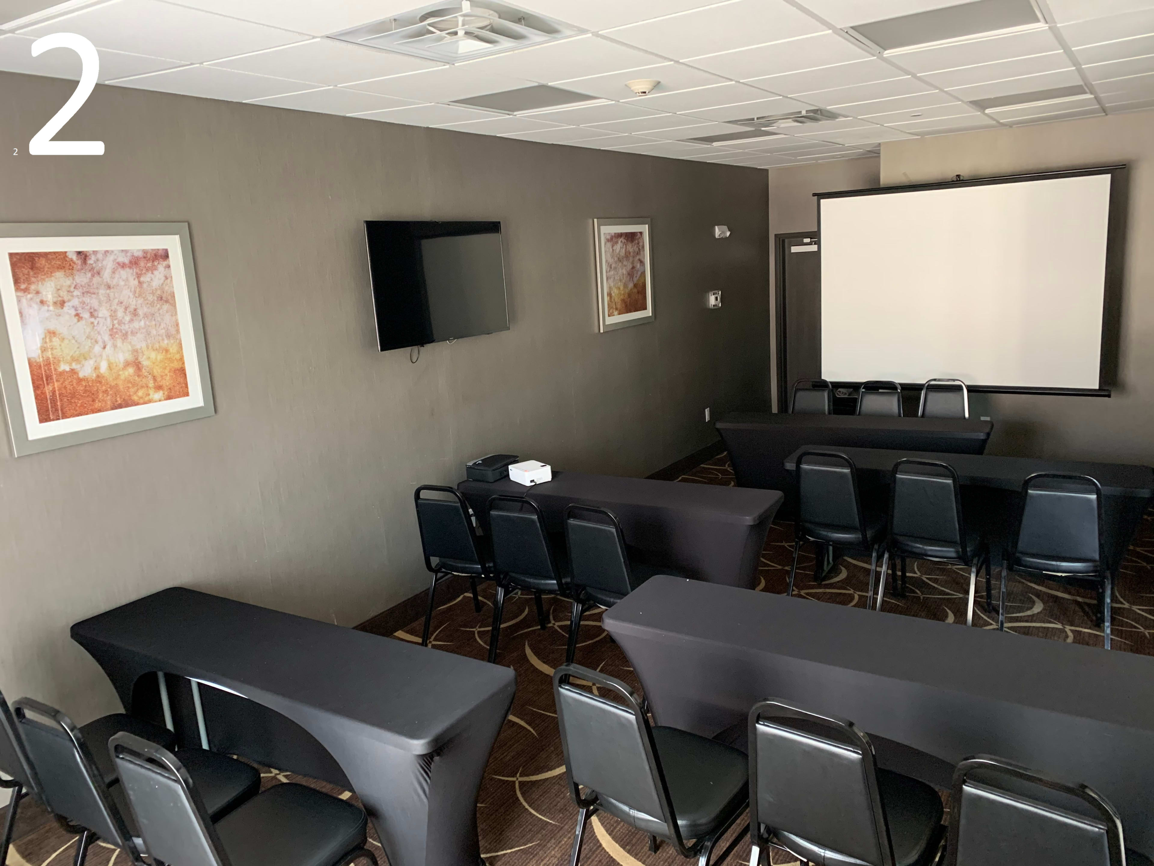 A small conference room featuring black tables and chairs, a projector screen, and a television mounted on the wall.