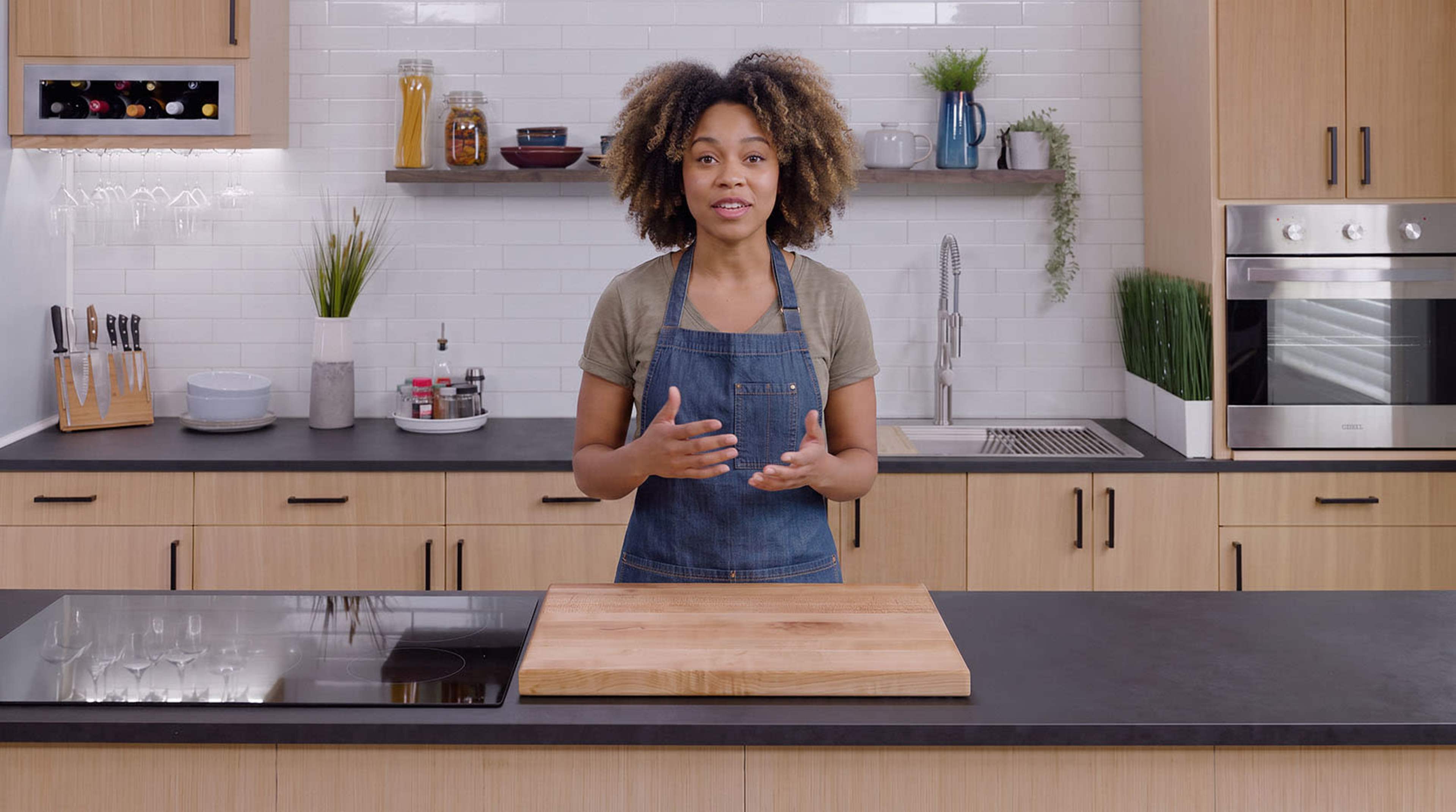 A person with curly hair stands in a modern kitchen, facing the camera with a wooden cutting board in front of them.