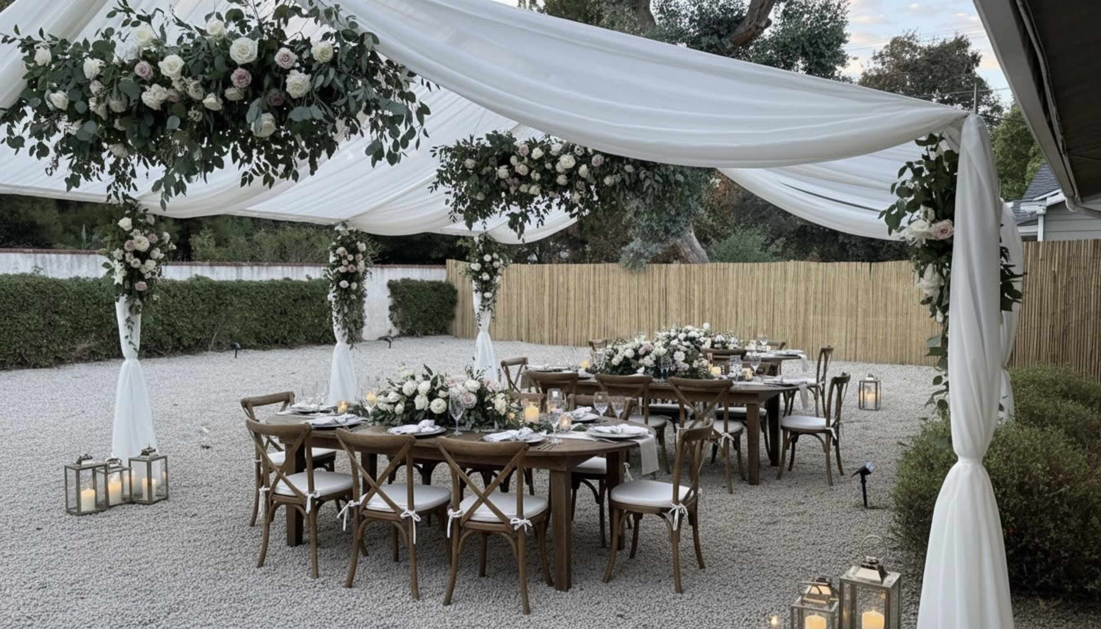 An outdoor dining setup features a long table adorned with centerpieces and is covered by a white canopy, surrounded by floral arrangements and lanterns.