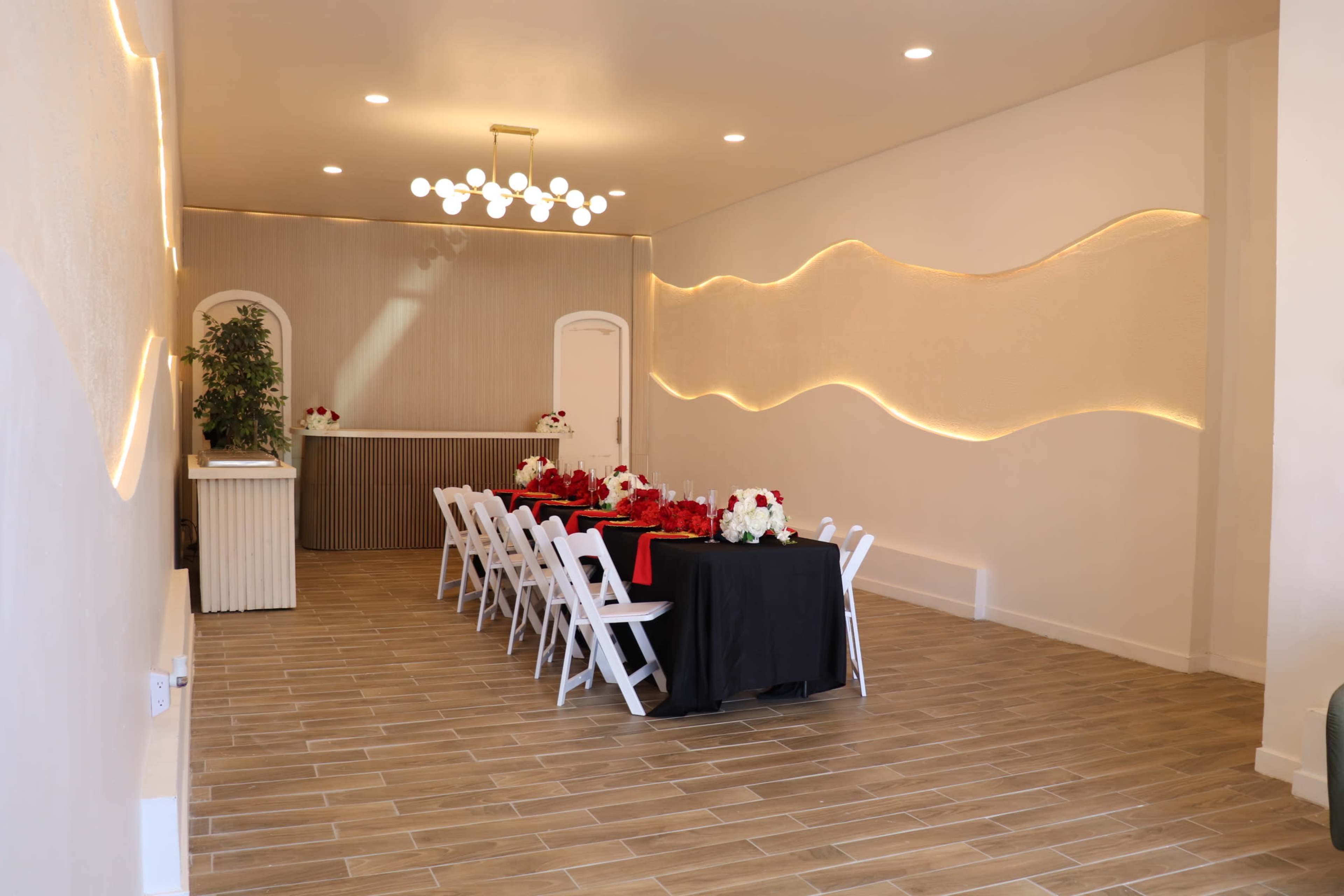 The image shows a modern event space with a long table set for a gathering, featuring a combination of white chairs and black tablecloths, accented by floral arrangements.