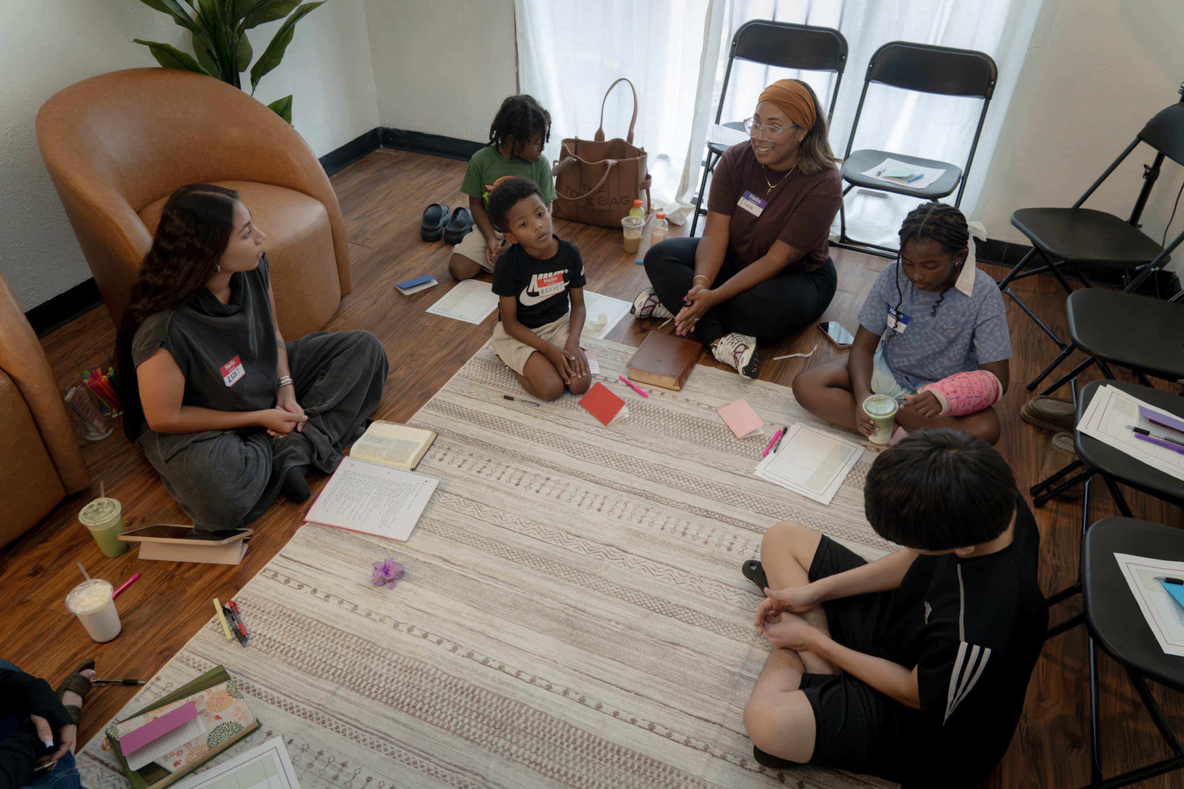 A diverse group of individuals is seated on the floor in a room, engaging in discussion while surrounded by notebooks and stationery.