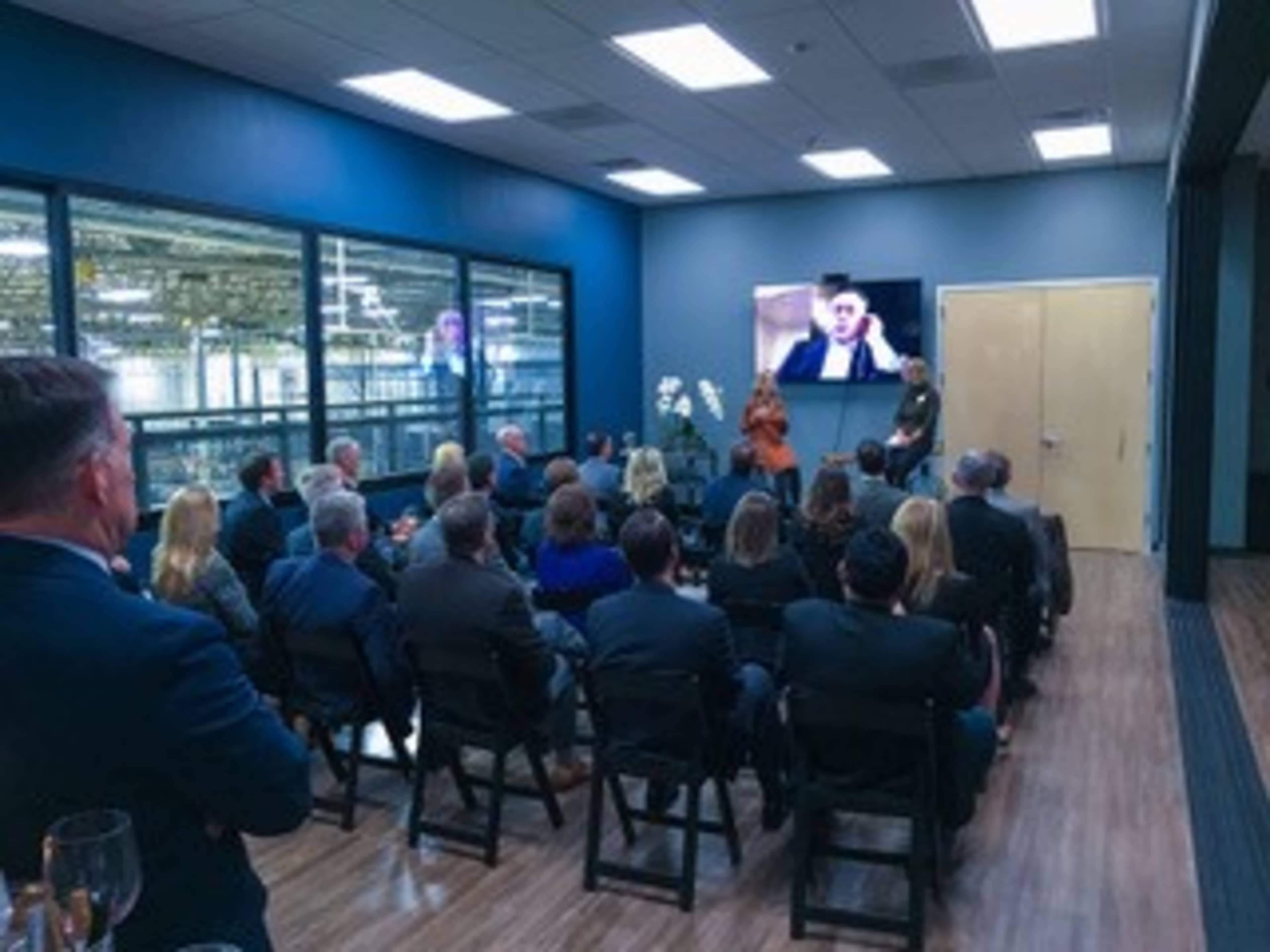 A group of people seated in a meeting room watches a presentation on a large screen while two speakers stand at the front.