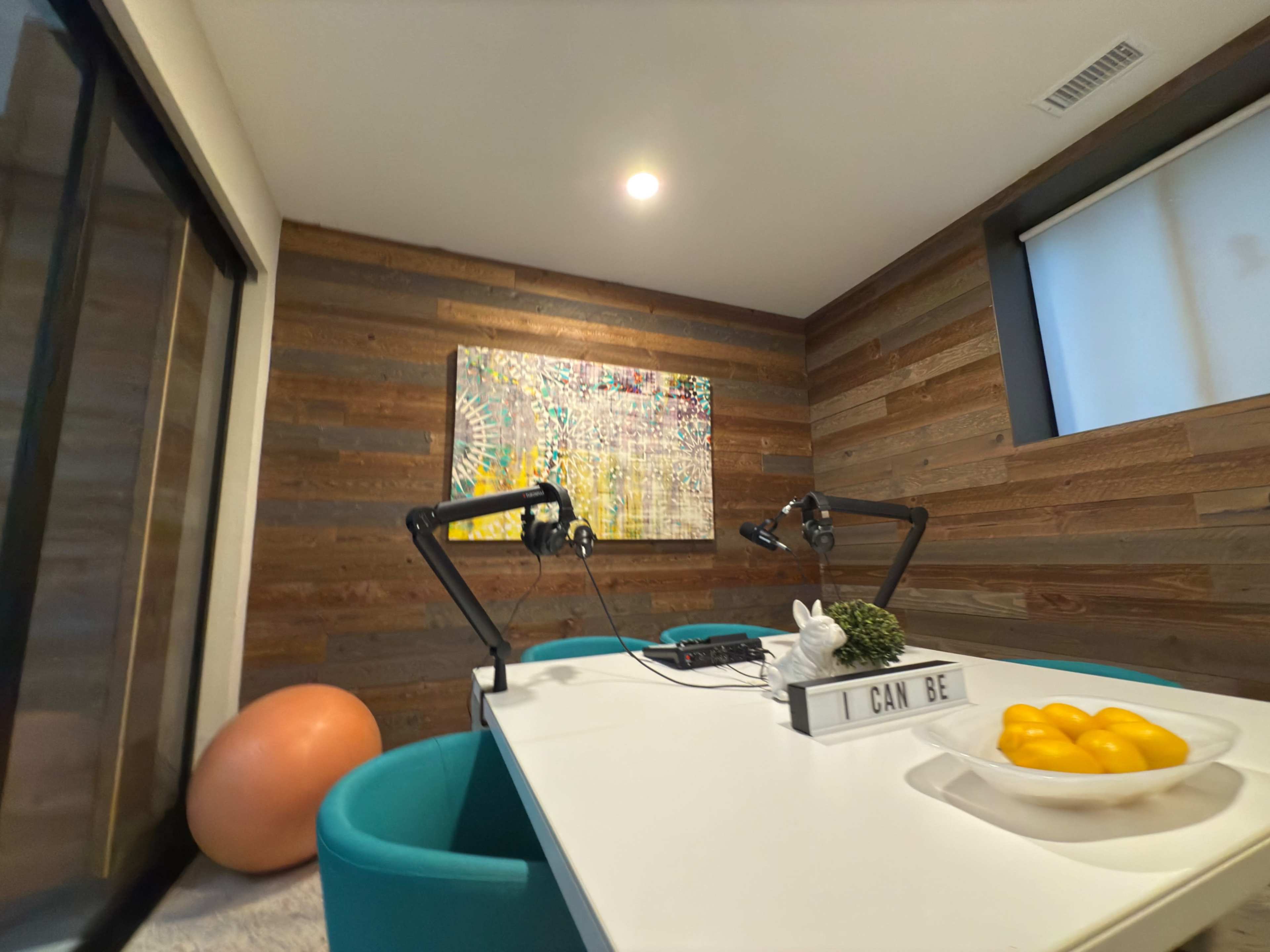 The image shows a modern workspace featuring a white table, turquoise chairs, a large piece of abstract art on the wall, and a decorative bowl of oranges.
