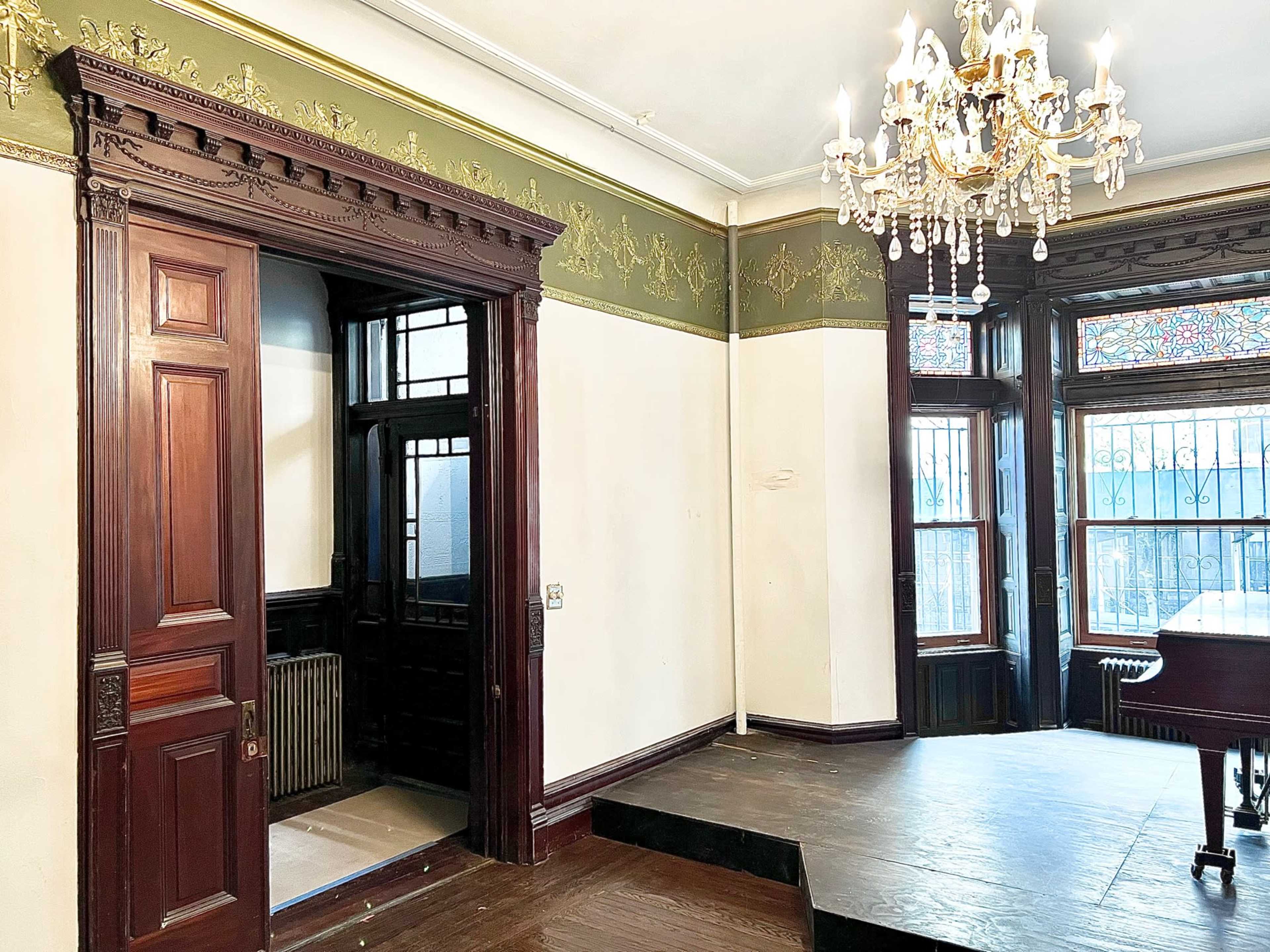 The image shows a spacious room featuring intricate woodwork, a chandelier, and a raised platform, with large windows that allow natural light to enter.