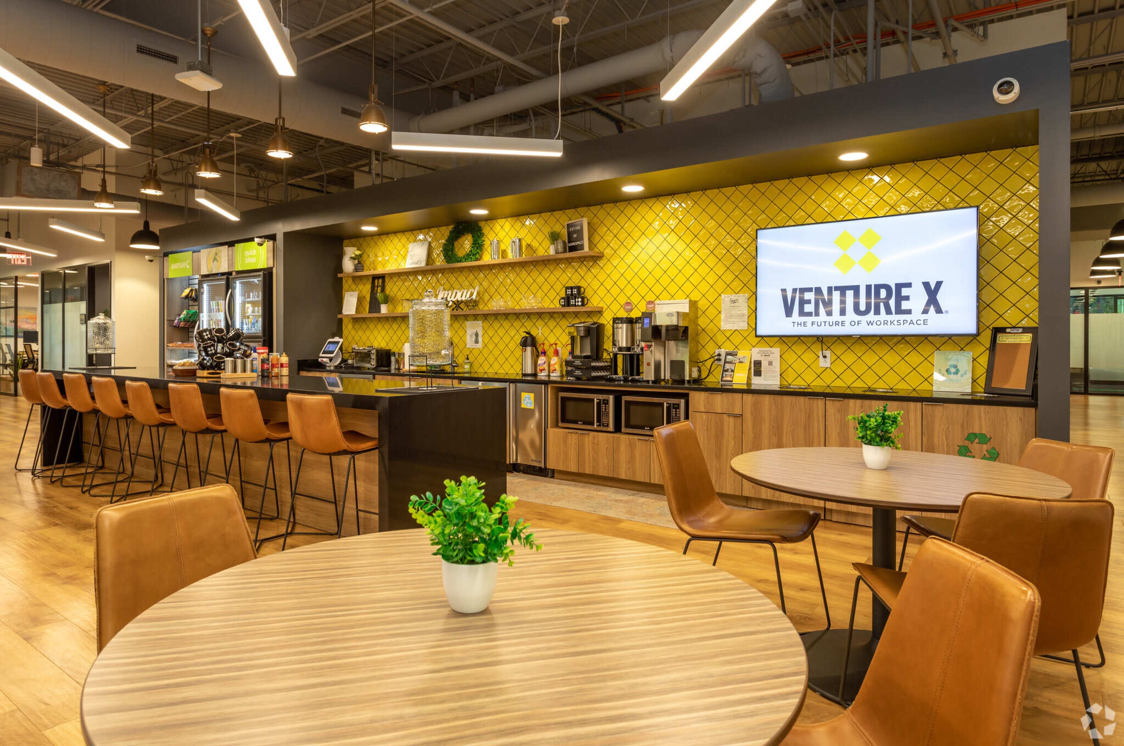 The image shows a modern coworking space featuring a bar area with yellow tiled walls, seating at a counter, and tables with plants.