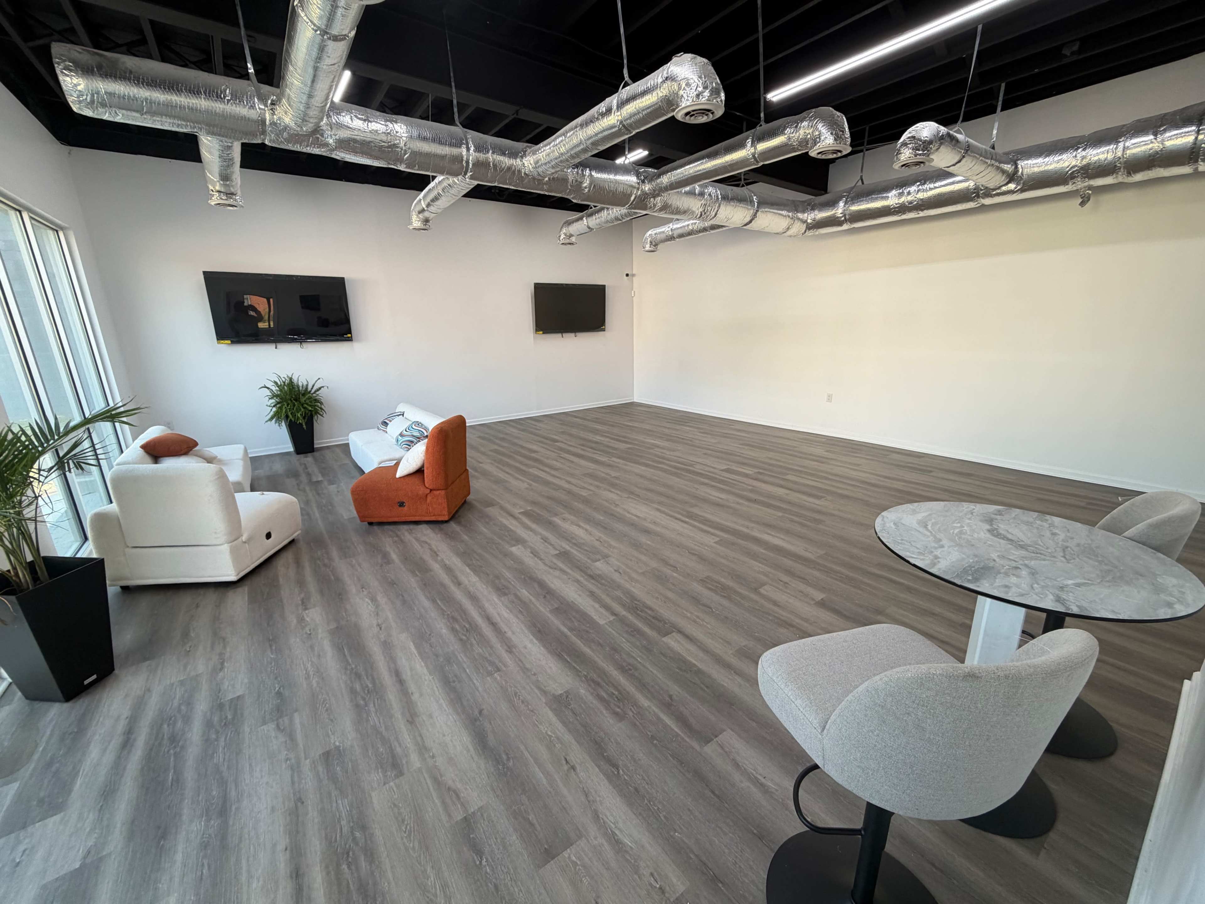 The image shows a spacious, modern room with wood-like flooring, featuring minimal furniture including a white couch, an orange chair, a small dining table with two chairs, and two wall-mounted screens.