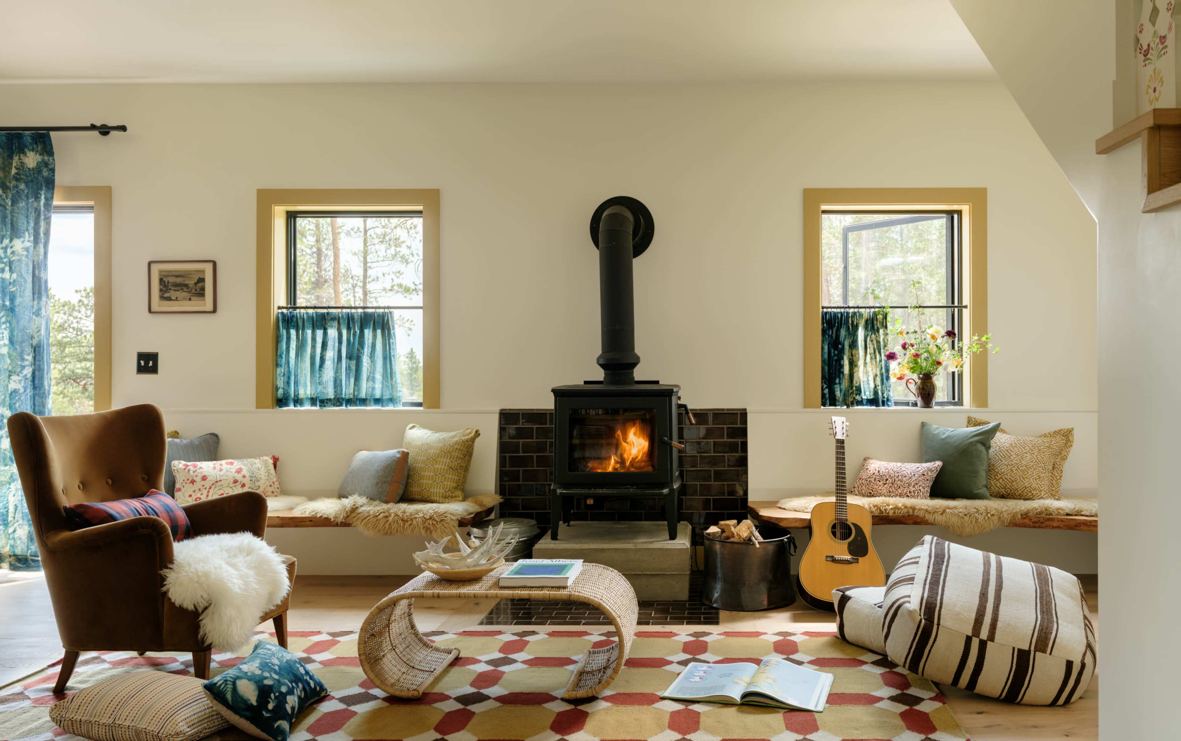 A cozy living room features a wood-burning stove, built-in seating with eclectic cushions, a guitar, and a colorful area rug.