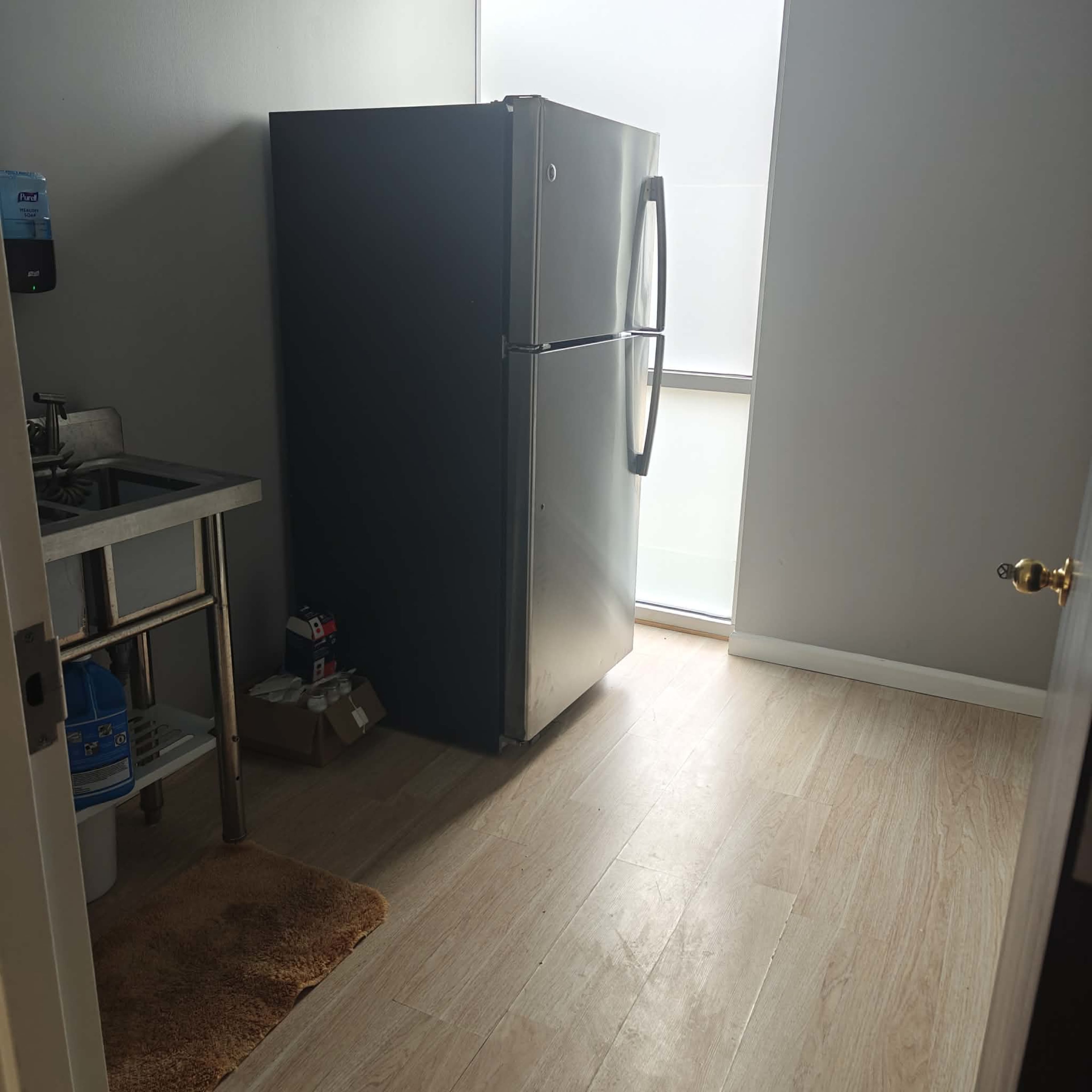 The image shows a kitchen corner with a large black refrigerator, a sink on the left, and light-colored wooden flooring.